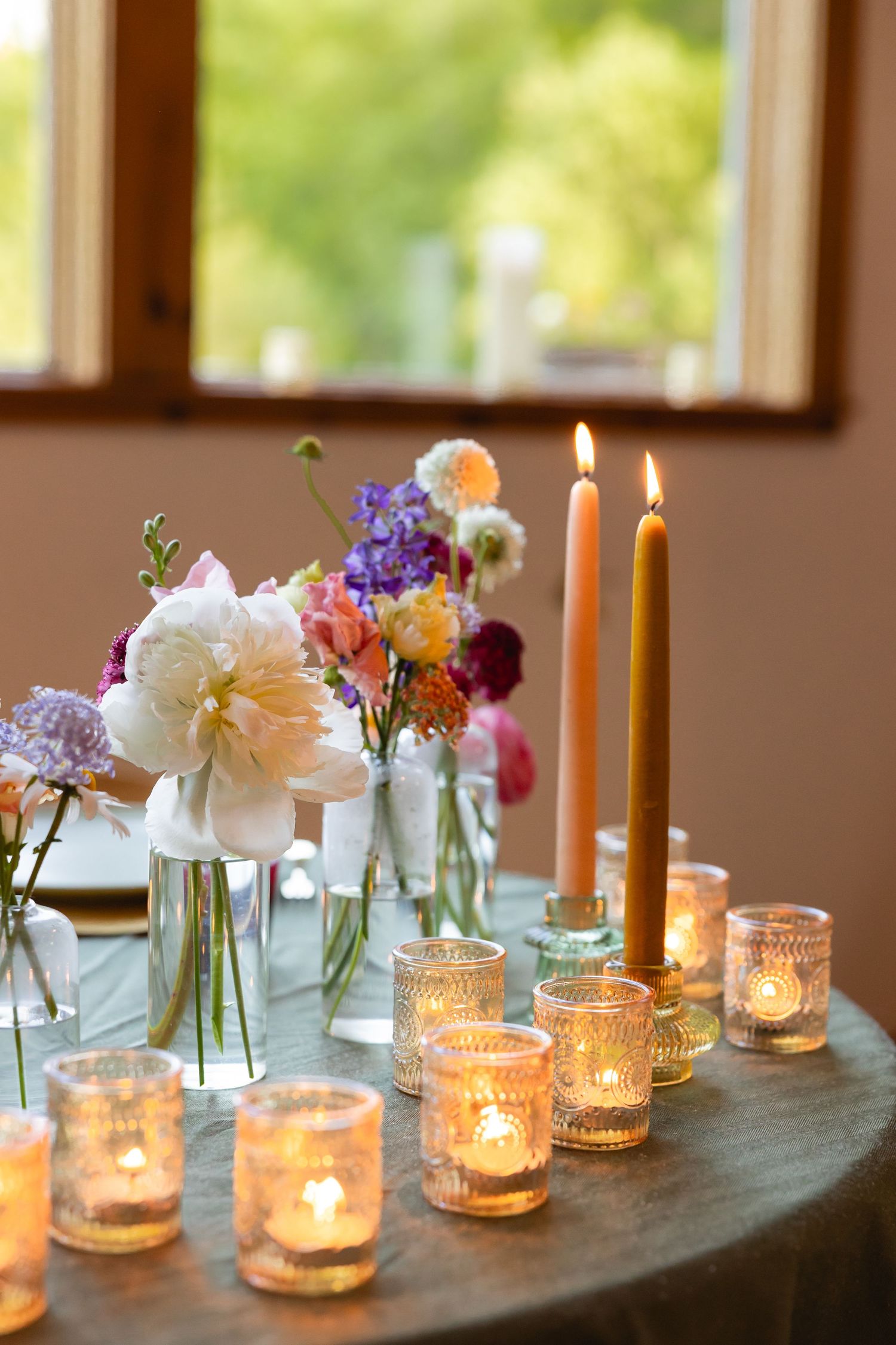 Row of glowing candles in glass holders alongside delicate flower arrangements on a gray tablecloth.
