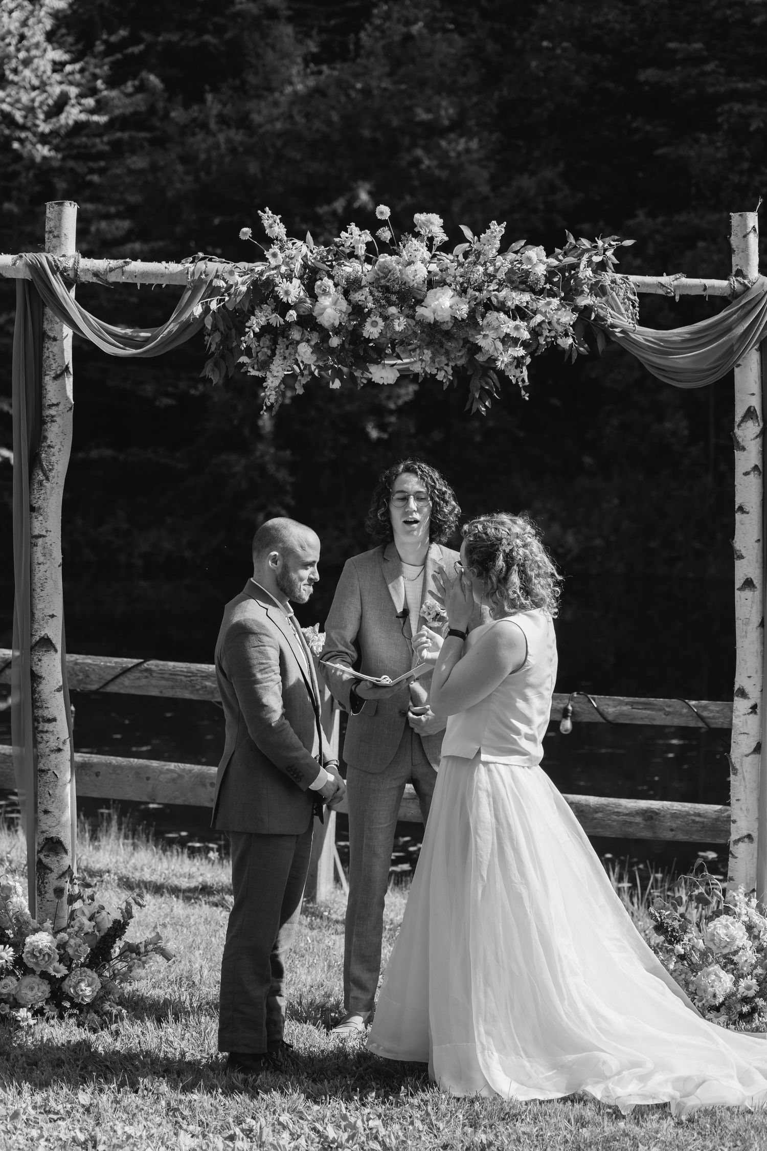 A wedding ceremony takes place under a rustic wooden arch adorned with white flowers and greenery in an outdoor setting.