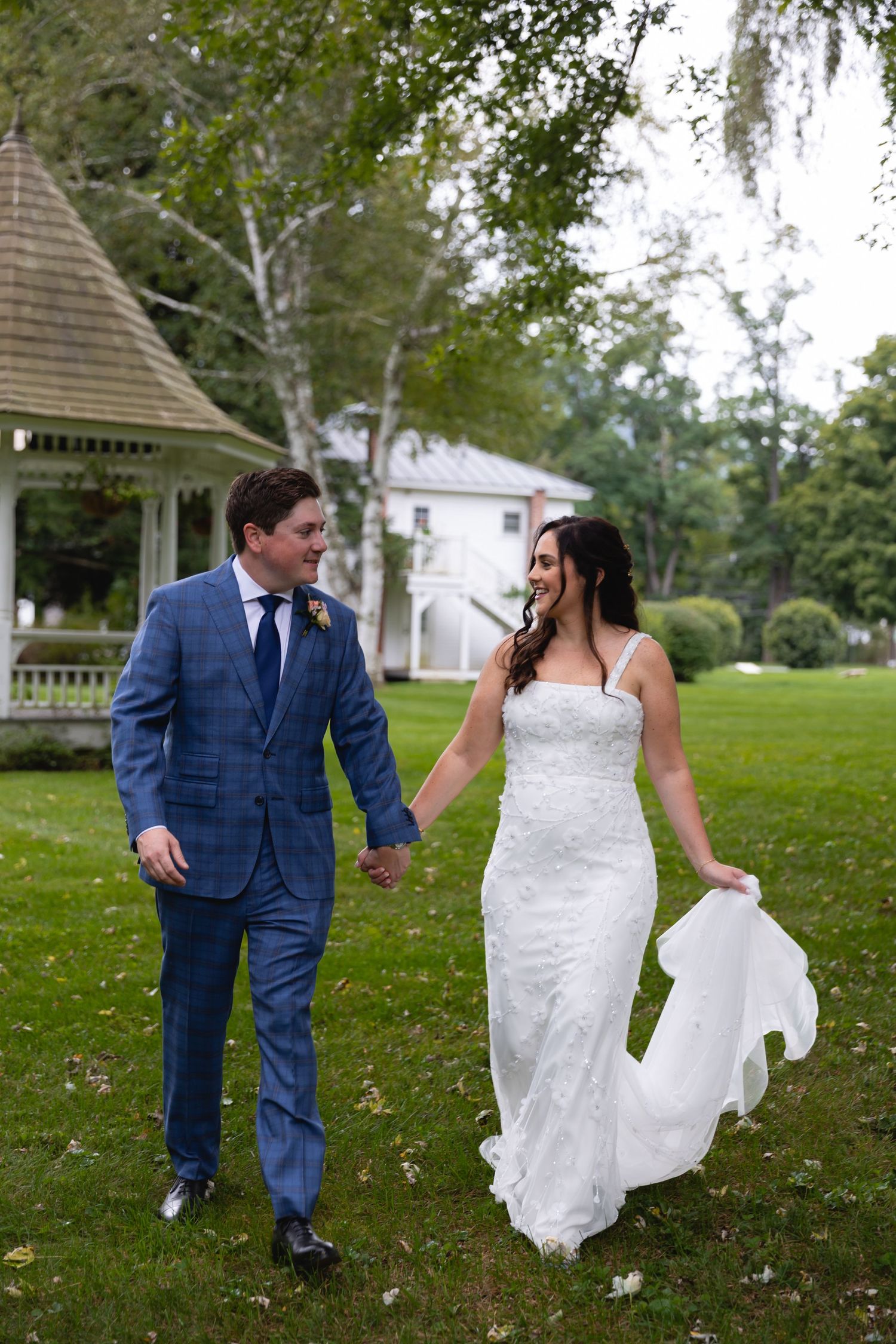 A couple in wedding attire walks together on a grass lawn with a gazebo and trees in the background.