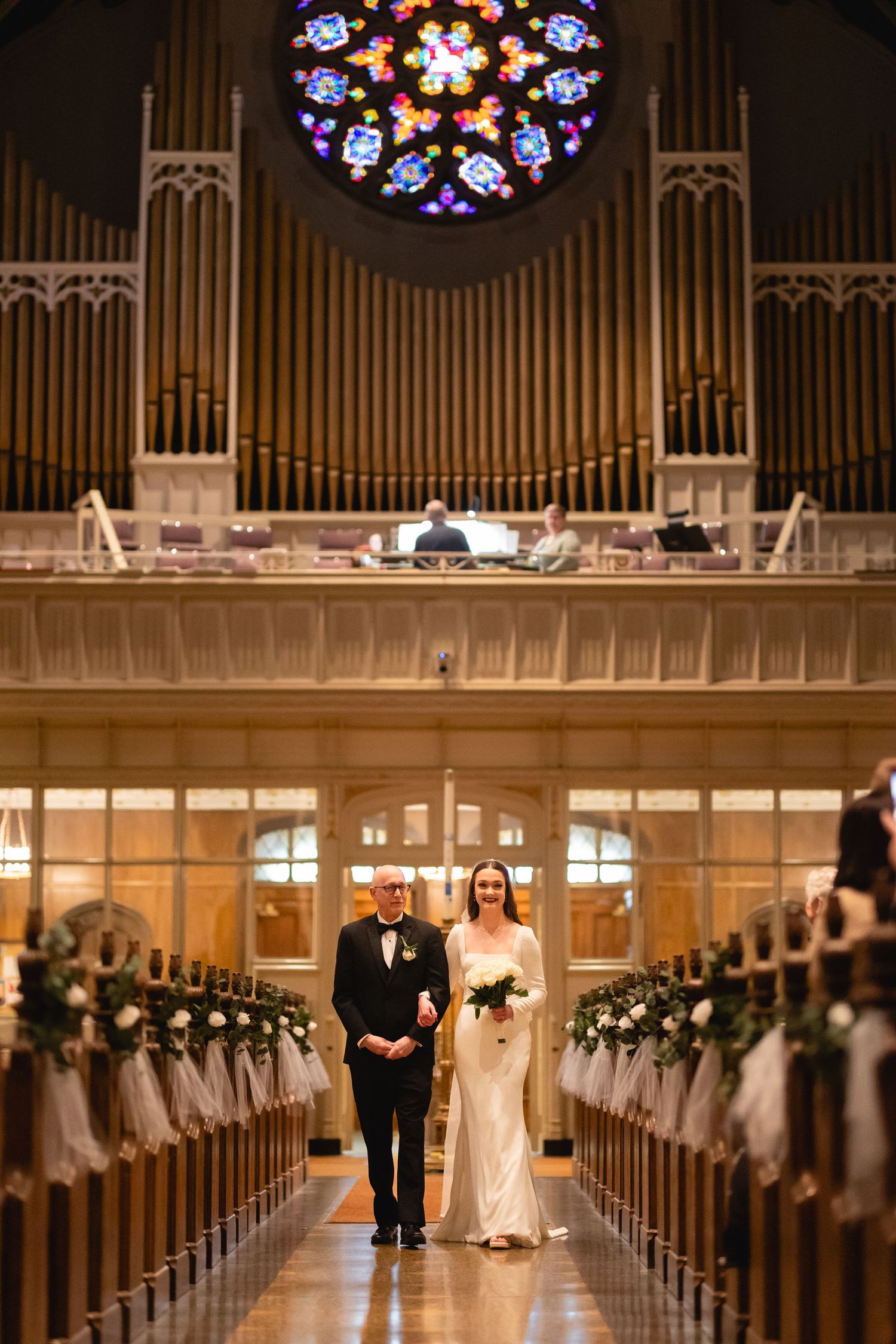 Elegant church ceremony with glowing stained glass window, pipe organ, and flower-decorated pews in grand cathedral setting.
