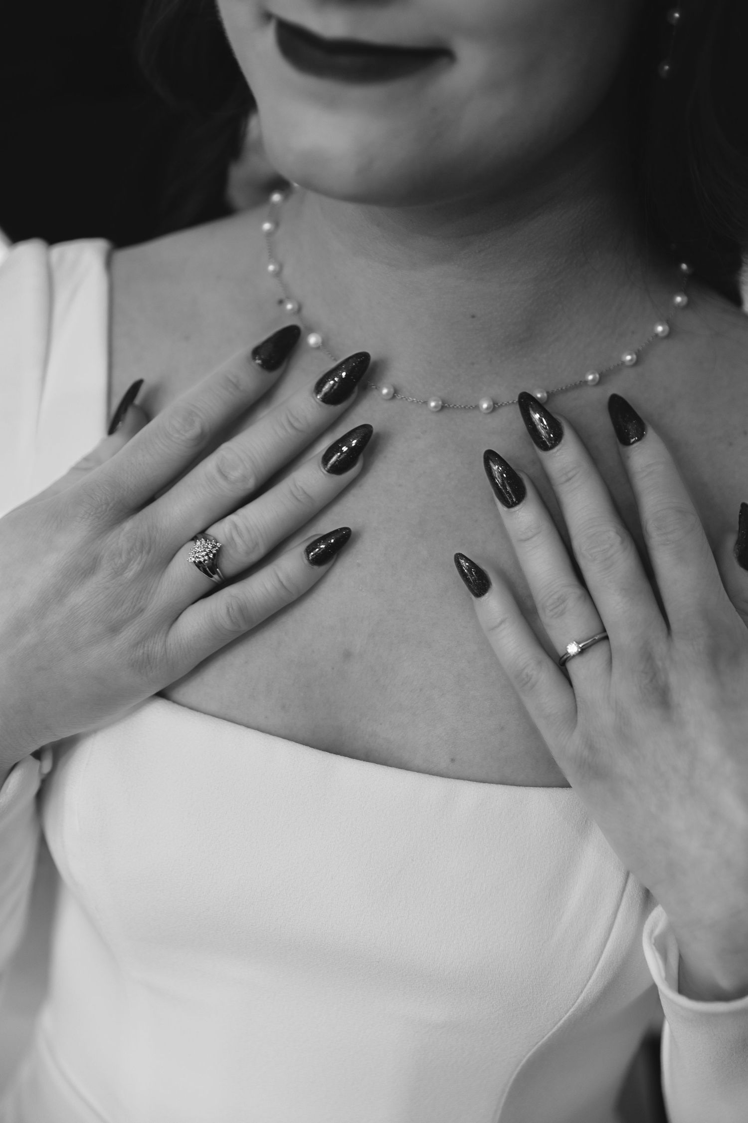 Black and white photo showing hands with dark manicured nails and delicate pearl necklace against white fabric.