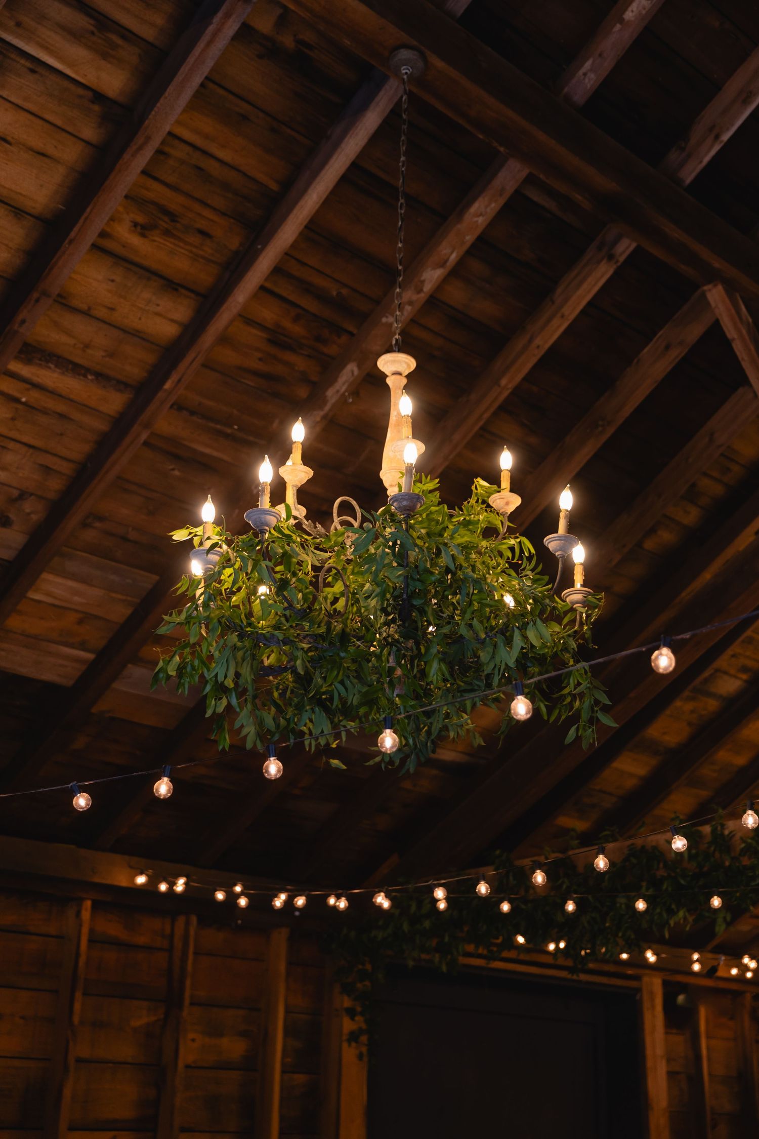 A rustic chandelier adorned with greenery and string lights hangs from wooden beams in a barn-style venue.