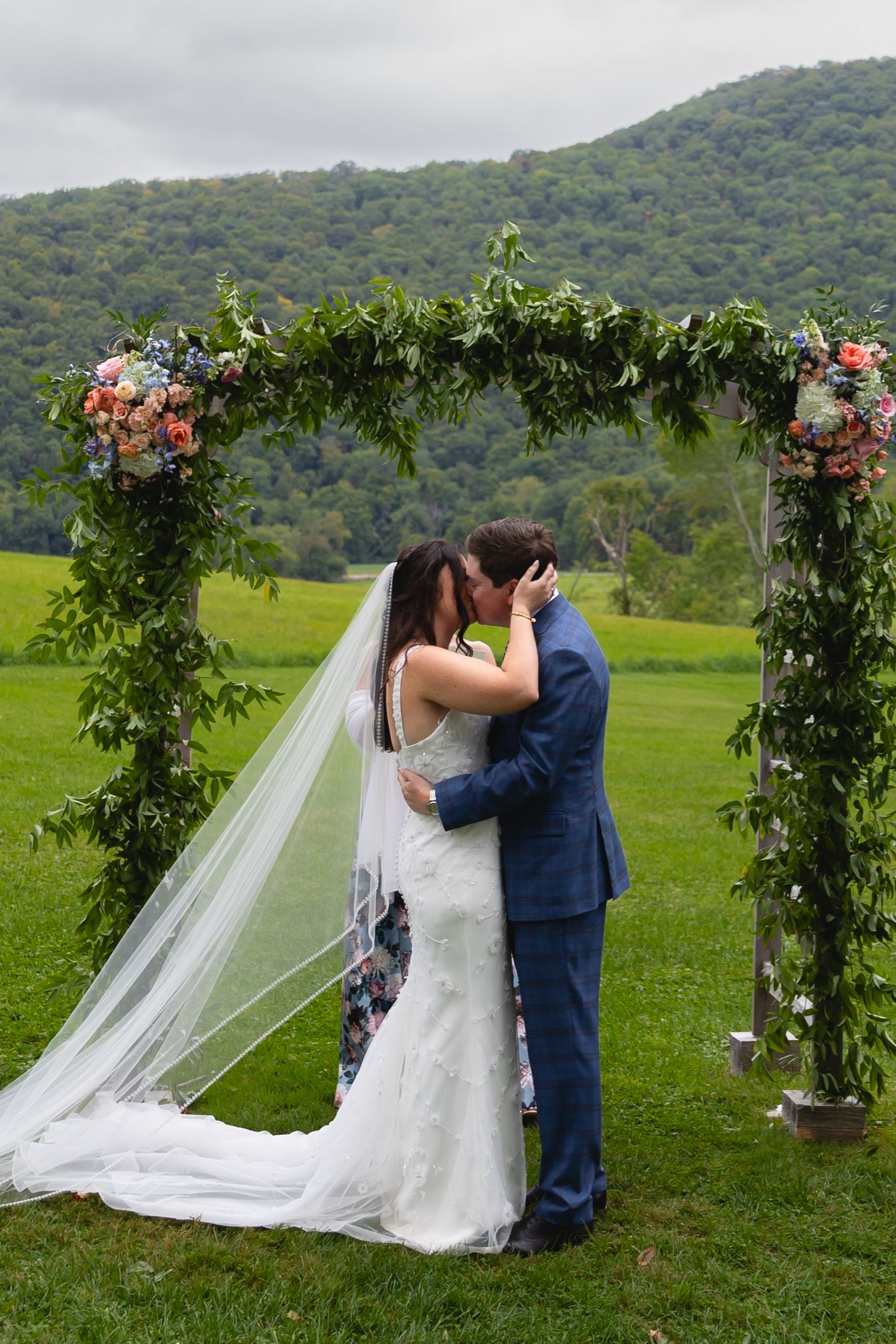A romantic wedding ceremony takes place under a floral arch on a lush green hillside with mountains in the background.