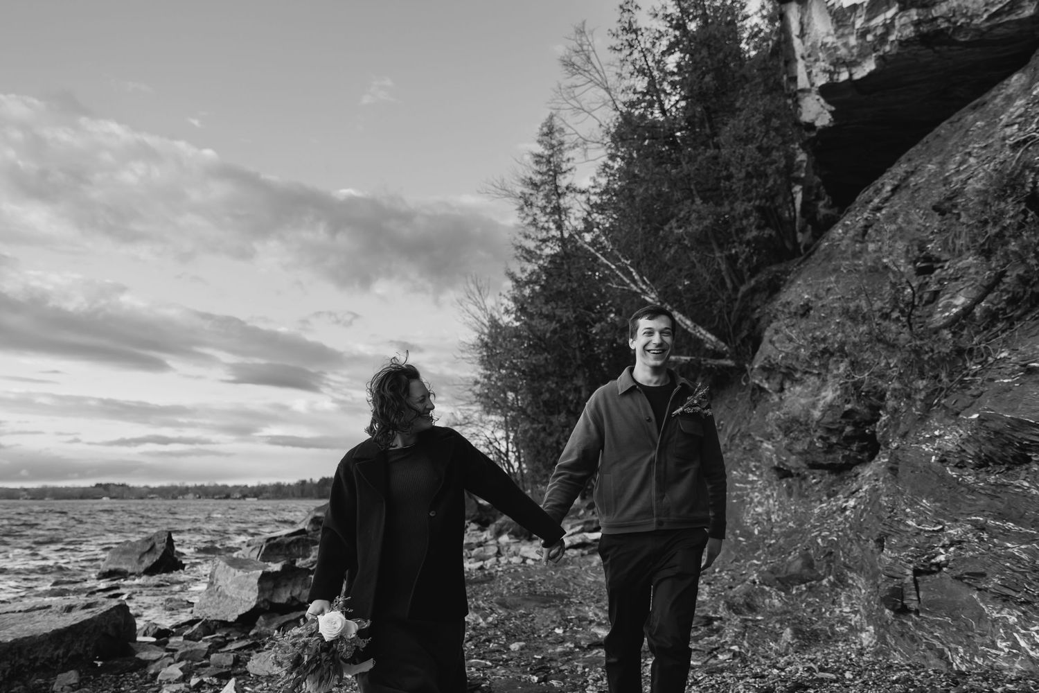 Two people holding hands while standing on rocky coastal cliffs with evergreen trees and dramatic clouds at sunset.