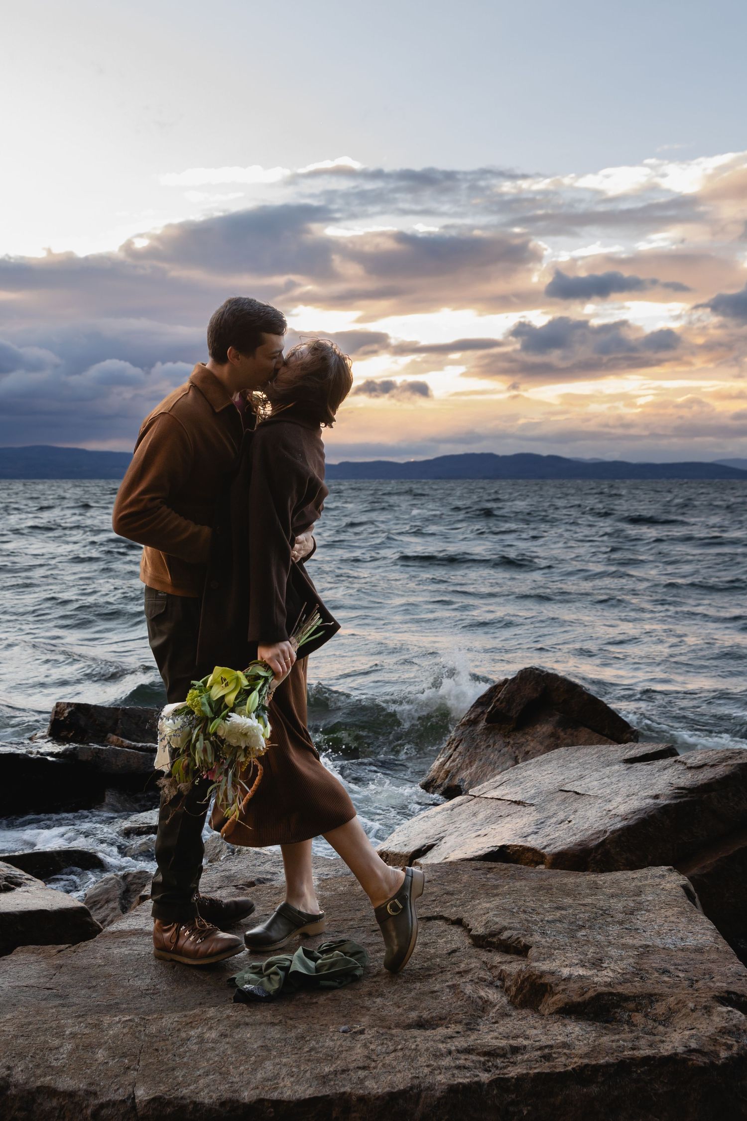 A romantic sunset moment on rocky coastal cliffs with dramatic cloudy skies and ocean waves crashing in the background.