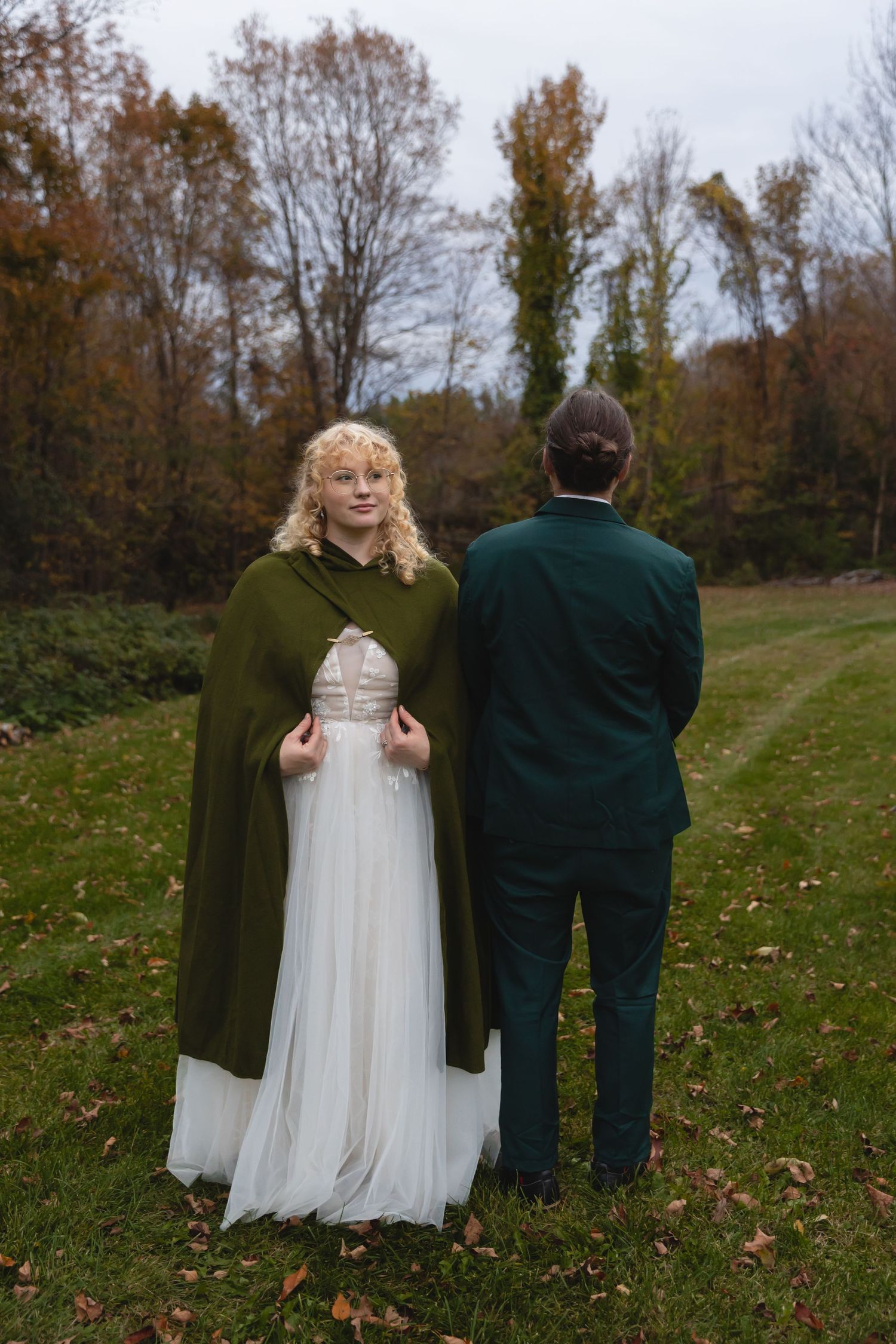 A bride in a white dress and green cape stands with her partner in an autumn field.