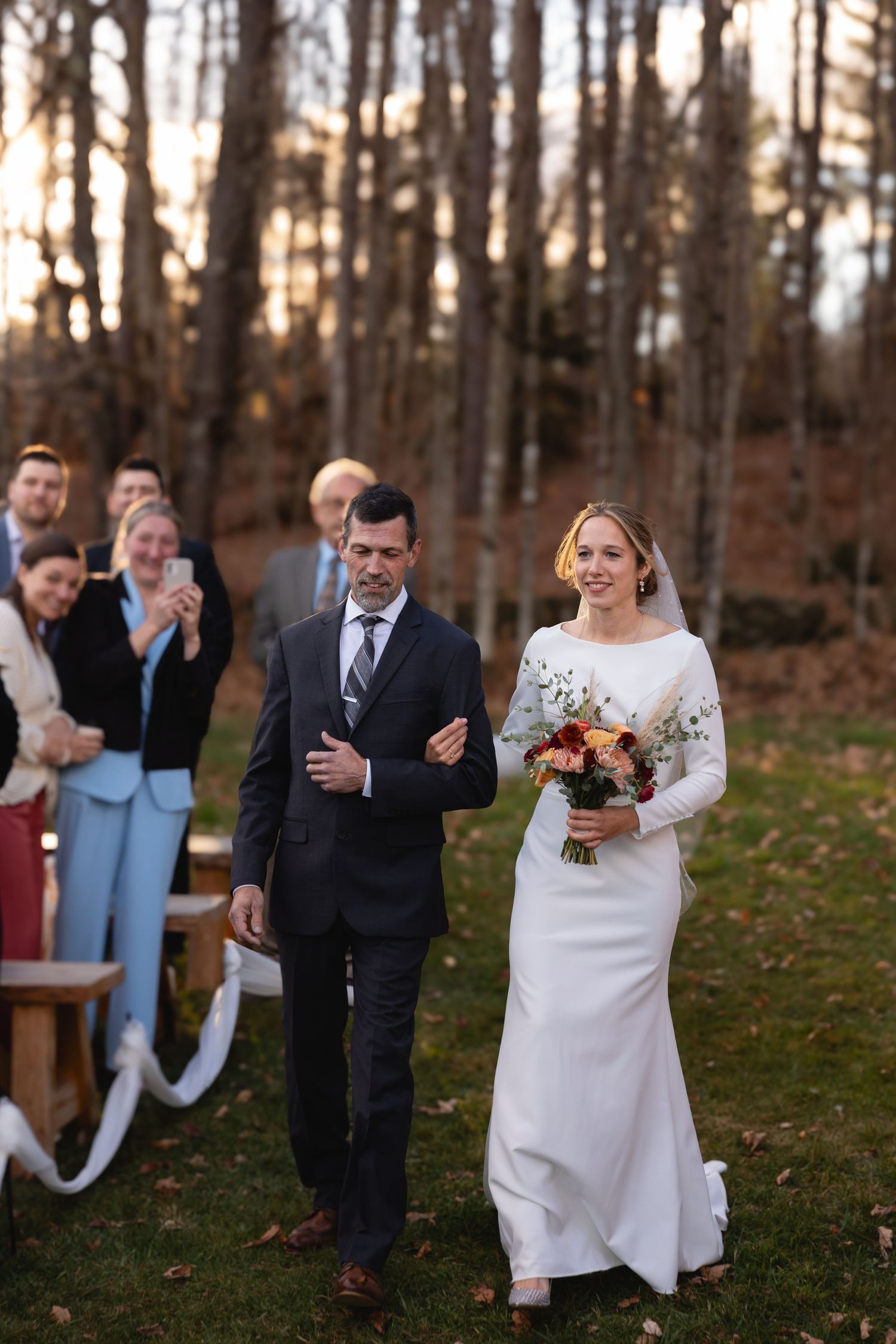 A wedding ceremony procession takes place in a wooded outdoor setting during autumn.