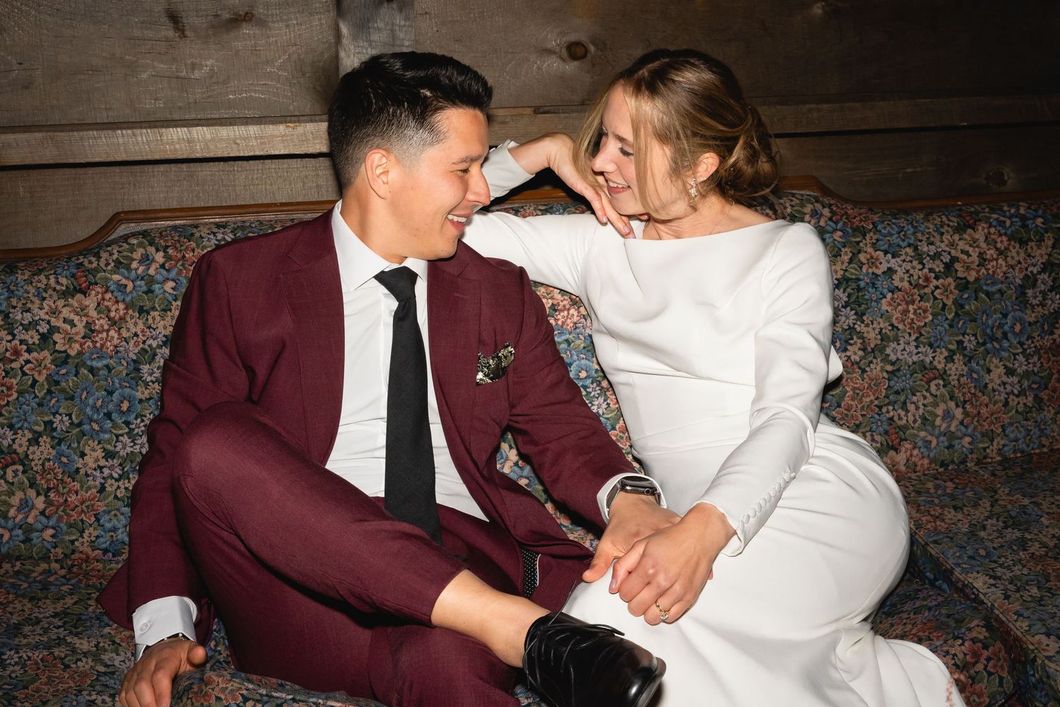 Happy couple laughs together on vintage sofa, bride in white dress and groom in burgundy suit.