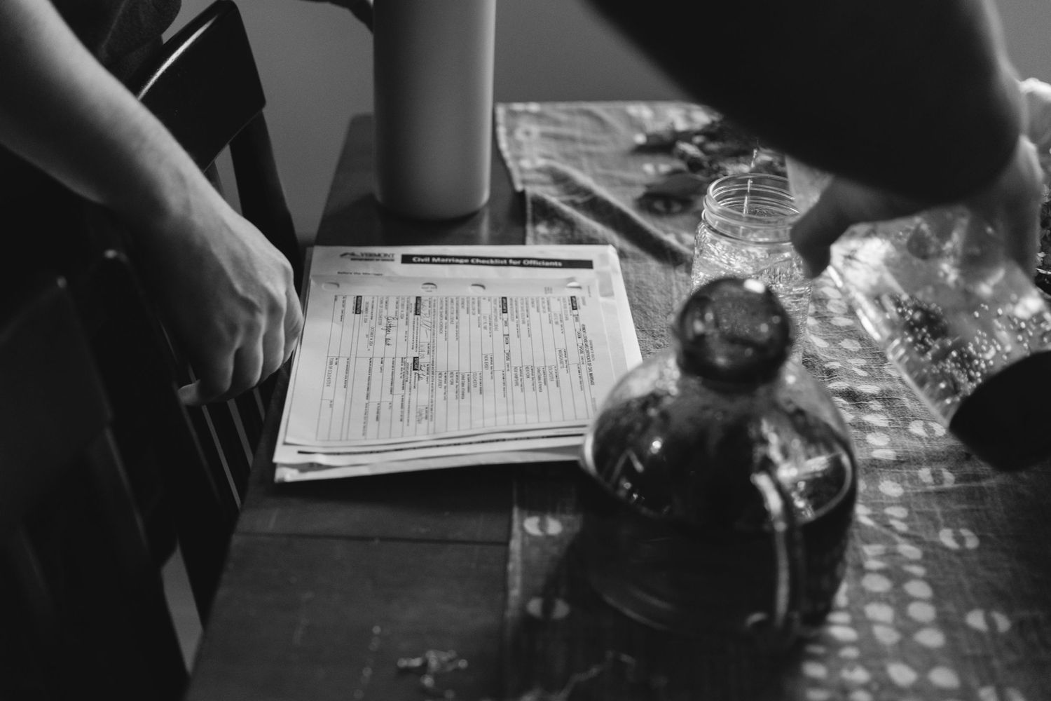 A person fills out a form on a table next to a glass bottle in black and white photography.