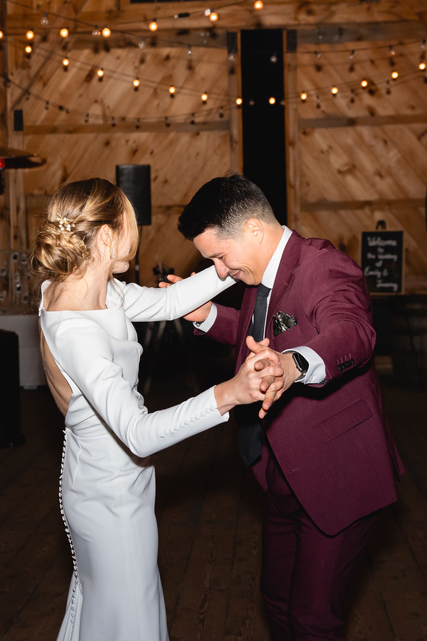 A bride and groom share their first dance together in a rustic barn venue with twinkling lights overhead.