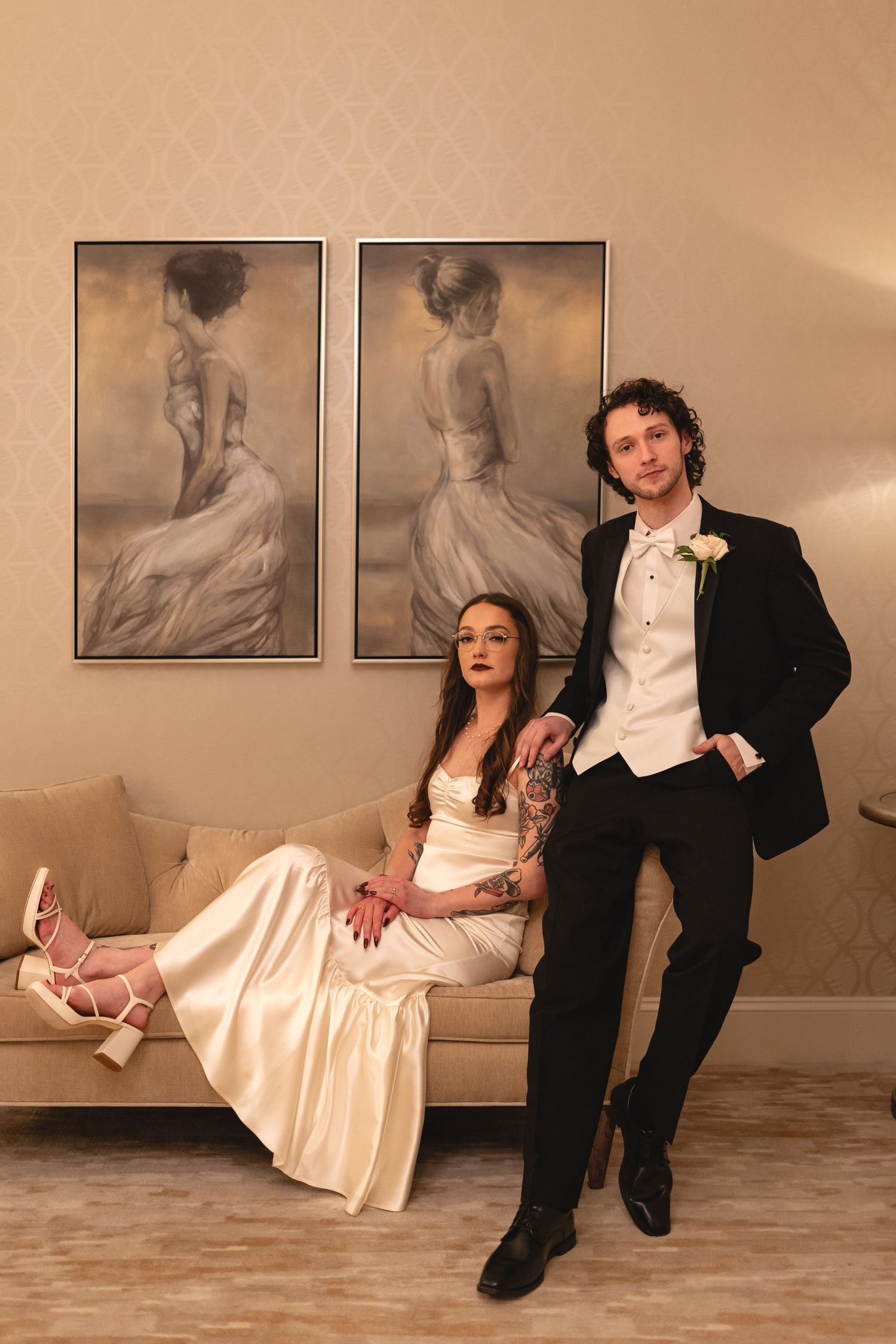 A couple in wedding attire poses against a wall with artistic portrait photographs in a warm-toned setting.