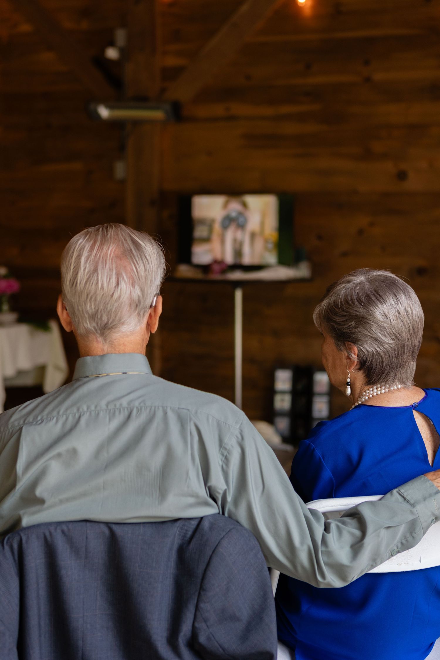 An elderly couple watches television together in a cozy room with wooden walls.