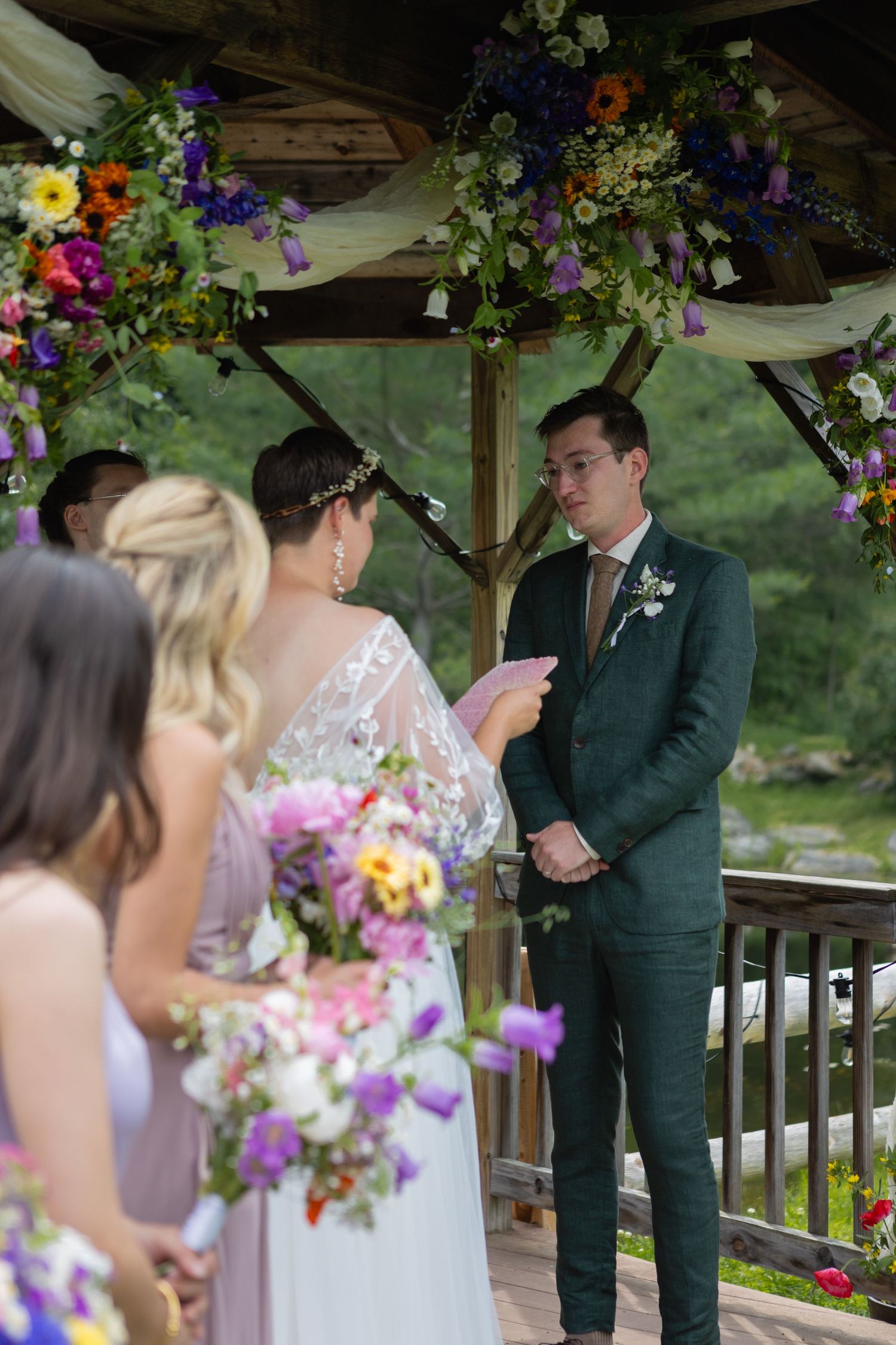 Wedding ceremony in progress under wooden gazebo with purple and orange floral decorations.