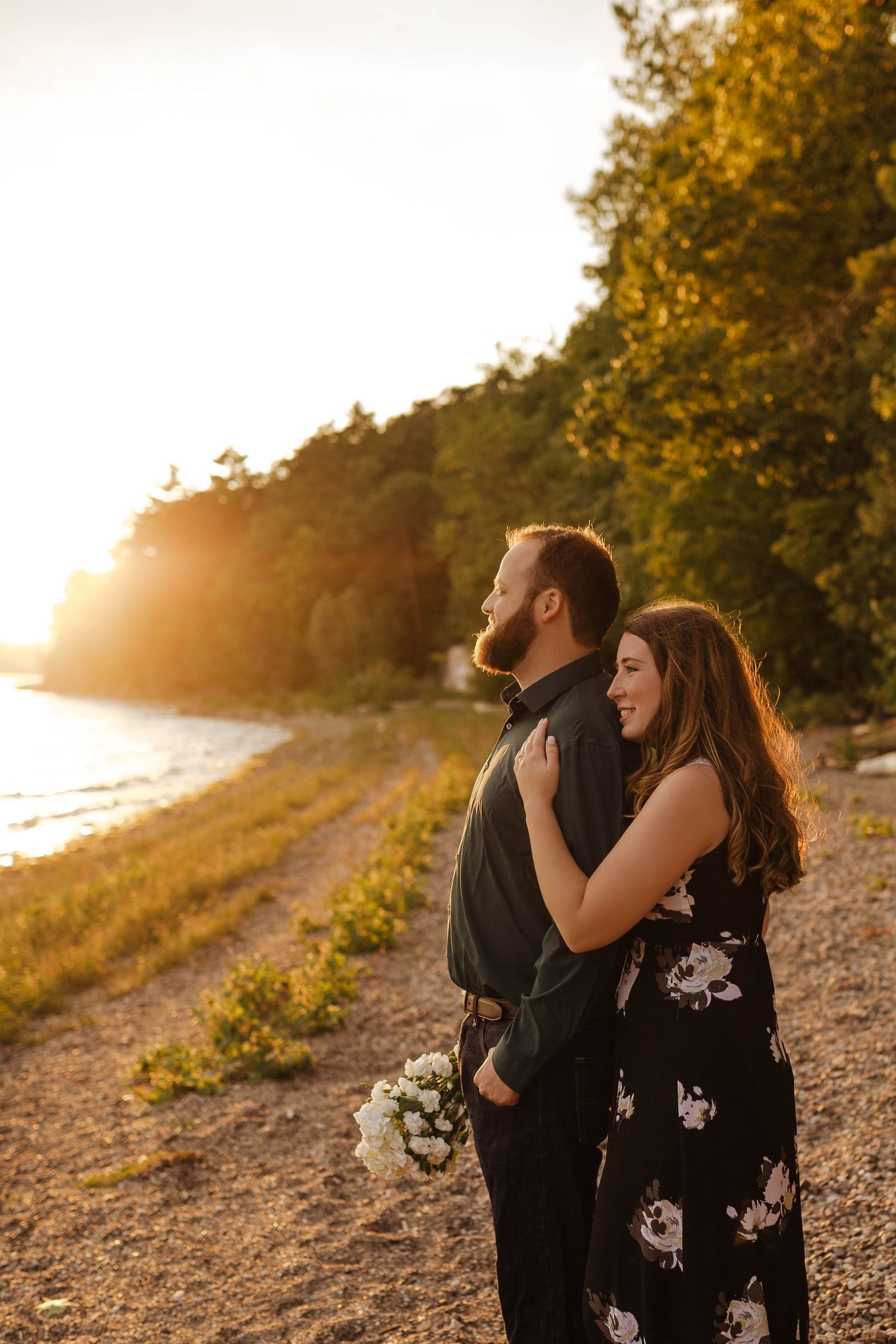 A couple embraces during golden hour on a scenic coastal path with flowers.