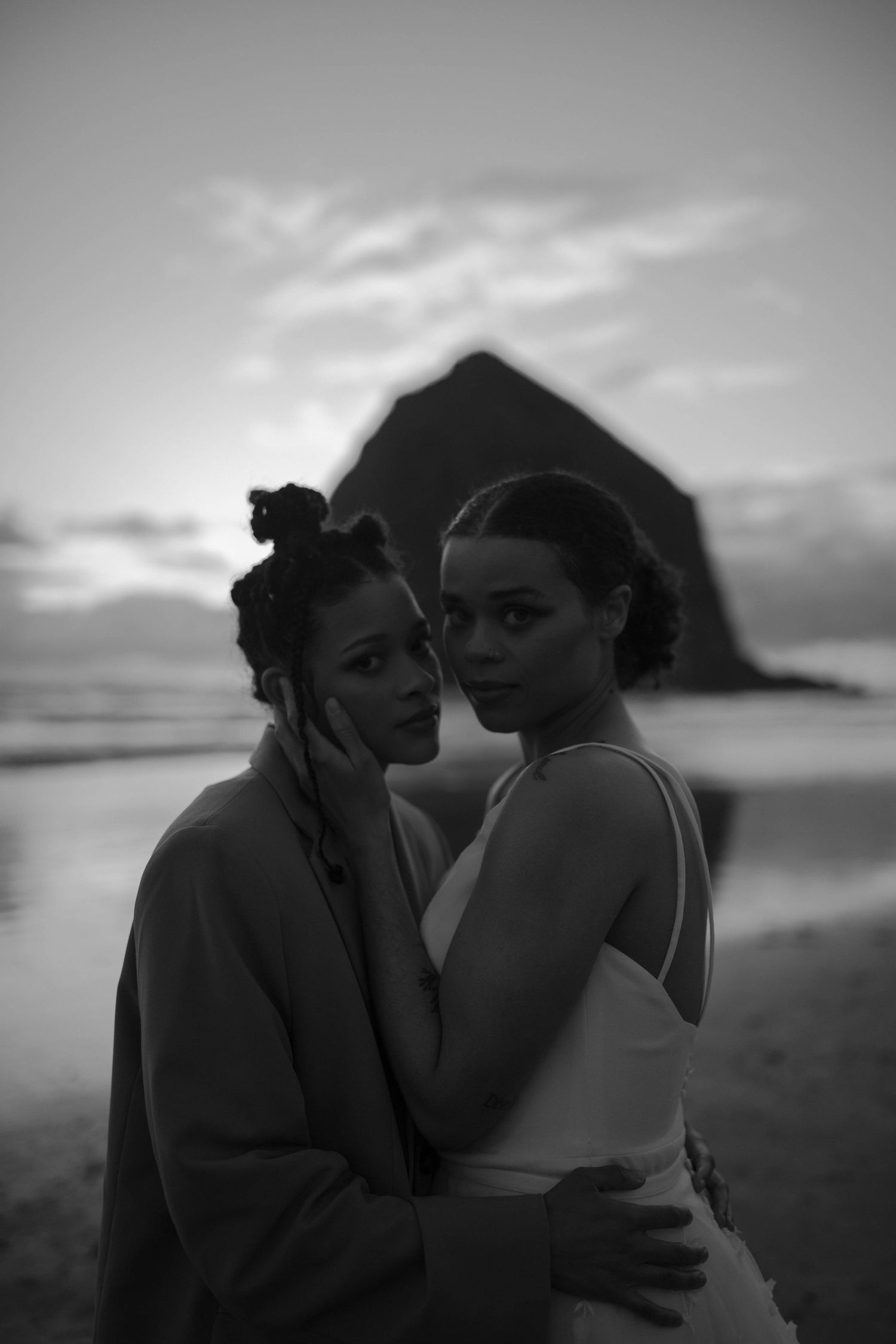 A black and white intimate portrait of newlyweds embracing on the beach with Haystack Rock visible.