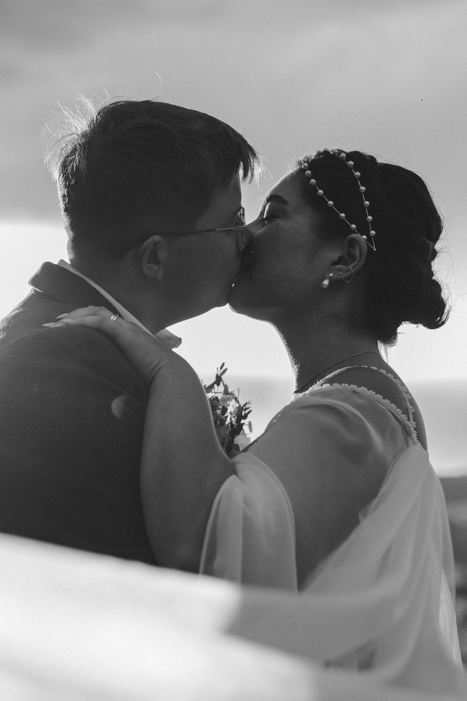 Black and white portrait of newlyweds sharing a romantic kiss against cloudy sky.
