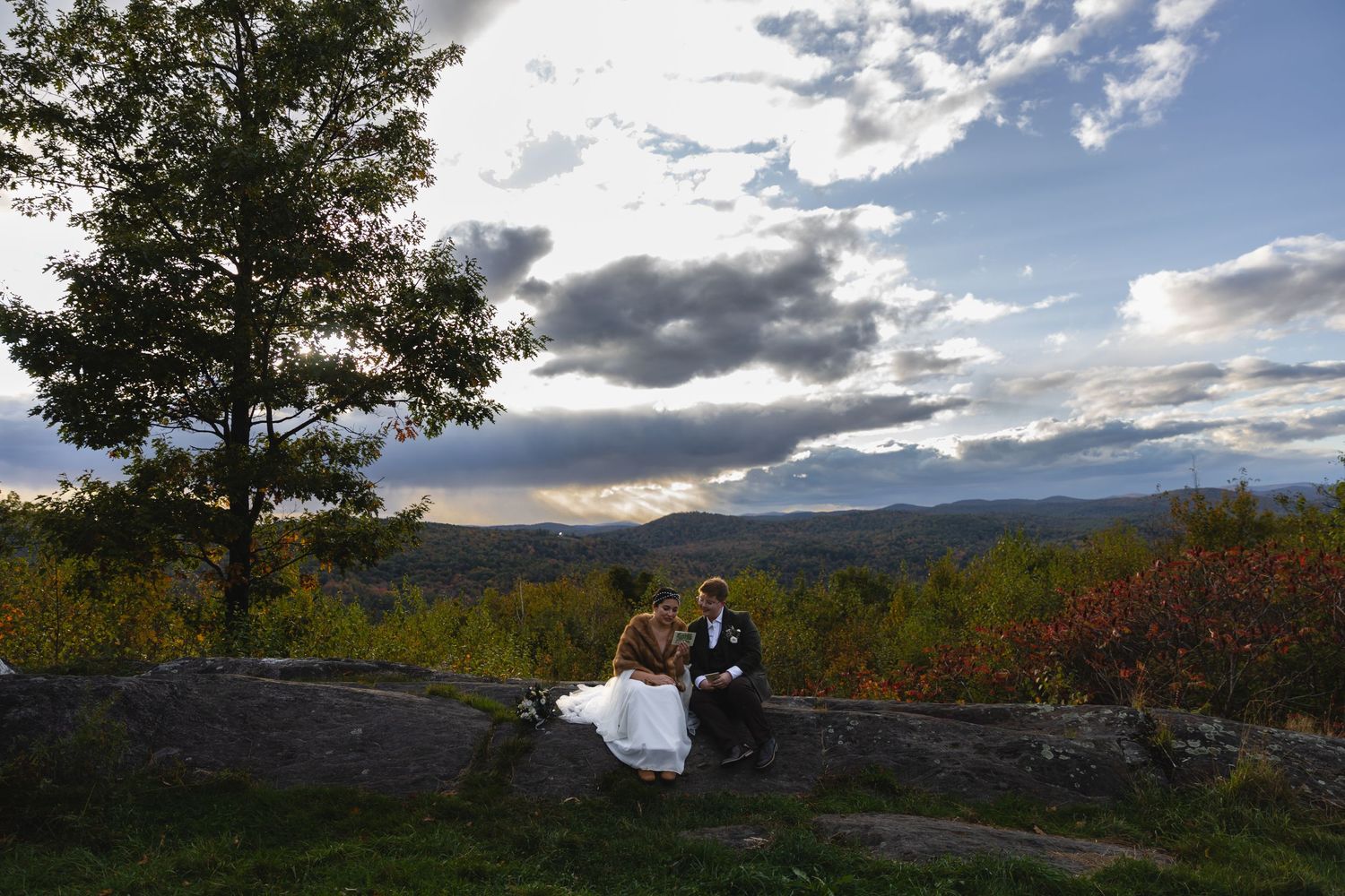 Wedding couple sitting on rocky outcrop with dramatic mountain vista and clouds behind them.
