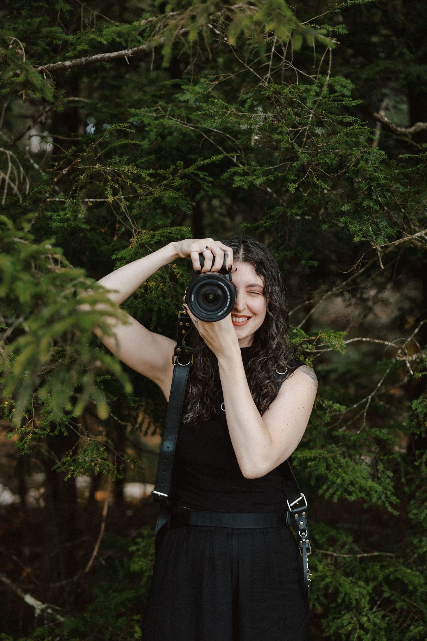 Smiling female photographer in black dress raises DSLR camera among lush green pine trees.