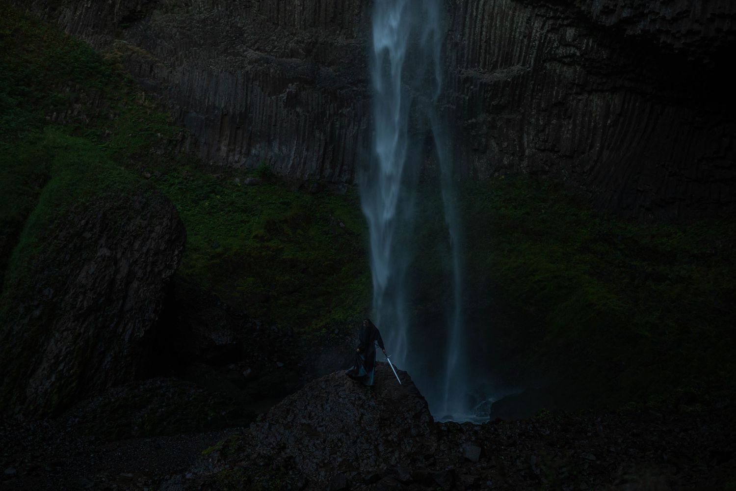 Tall waterfall cascades down dark rocky cliff face surrounded by green moss.