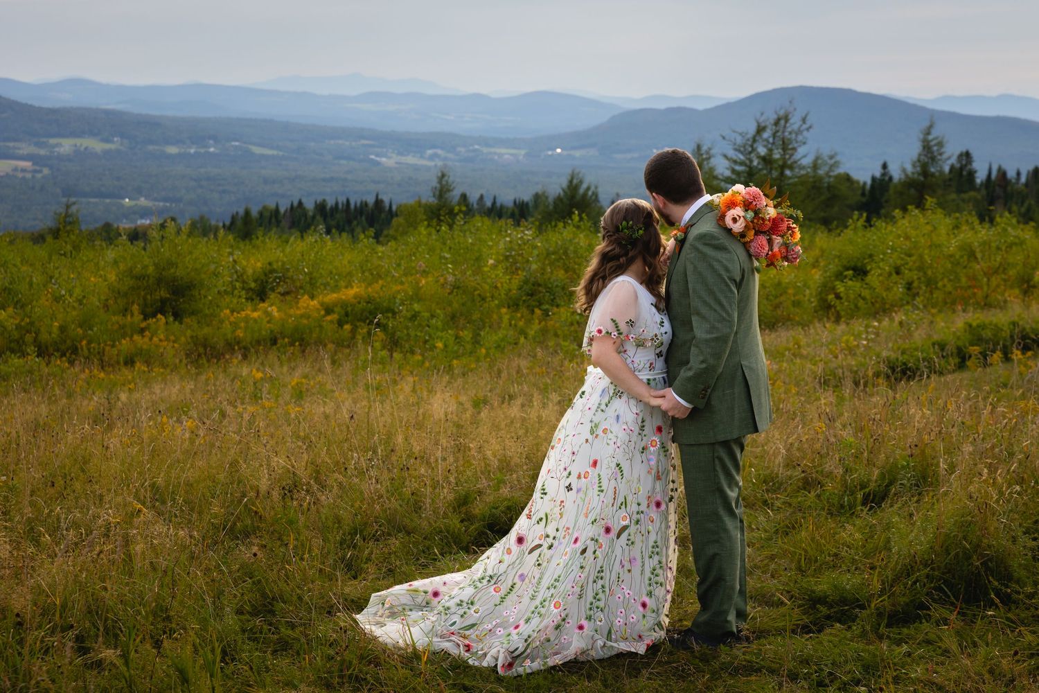A couple in wedding attire stands together on a scenic hilltop overlooking rolling mountains and green valleys.