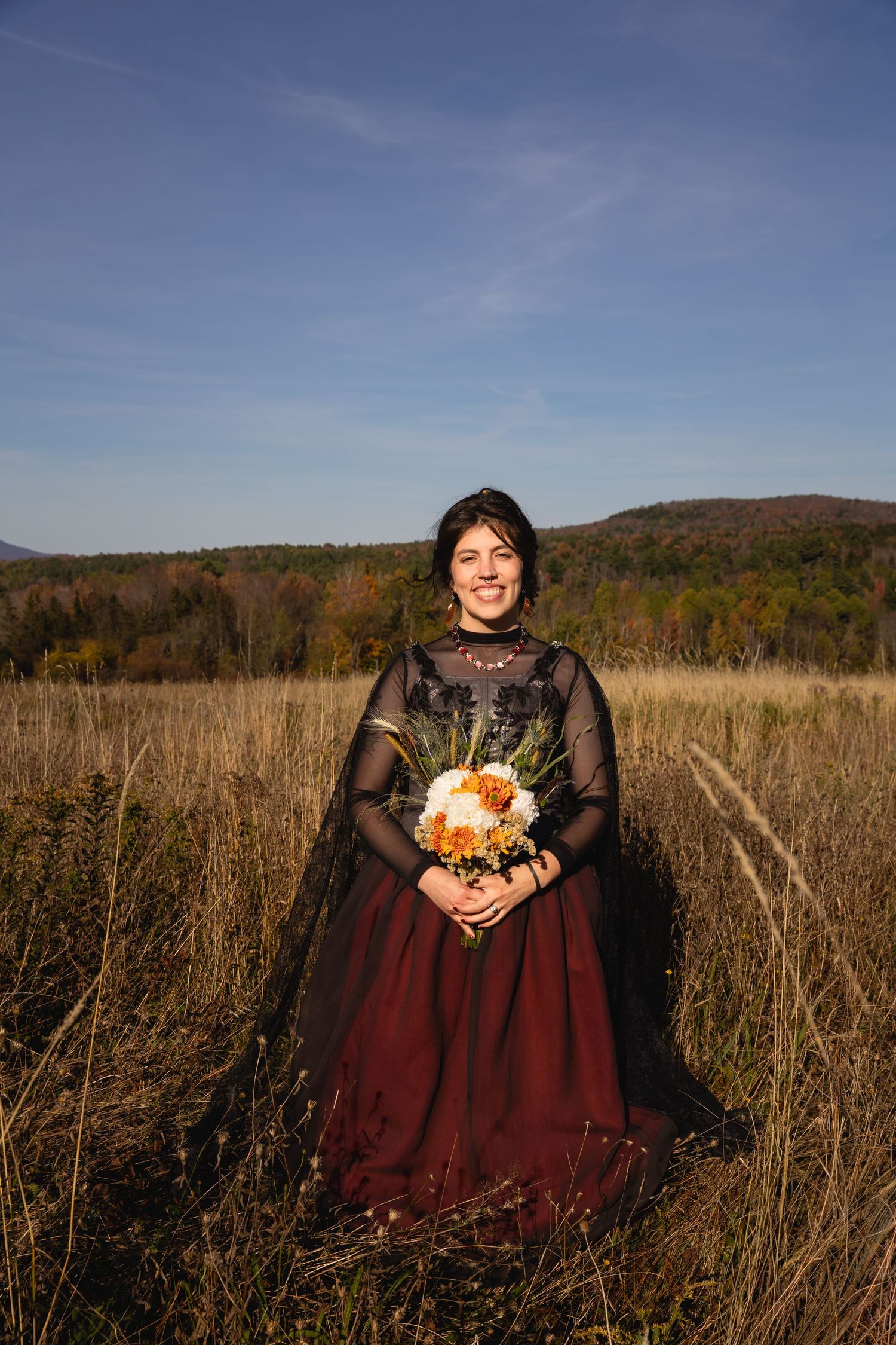 A figure in a burgundy dress holds an autumn bouquet while standing in a golden field at sunset.