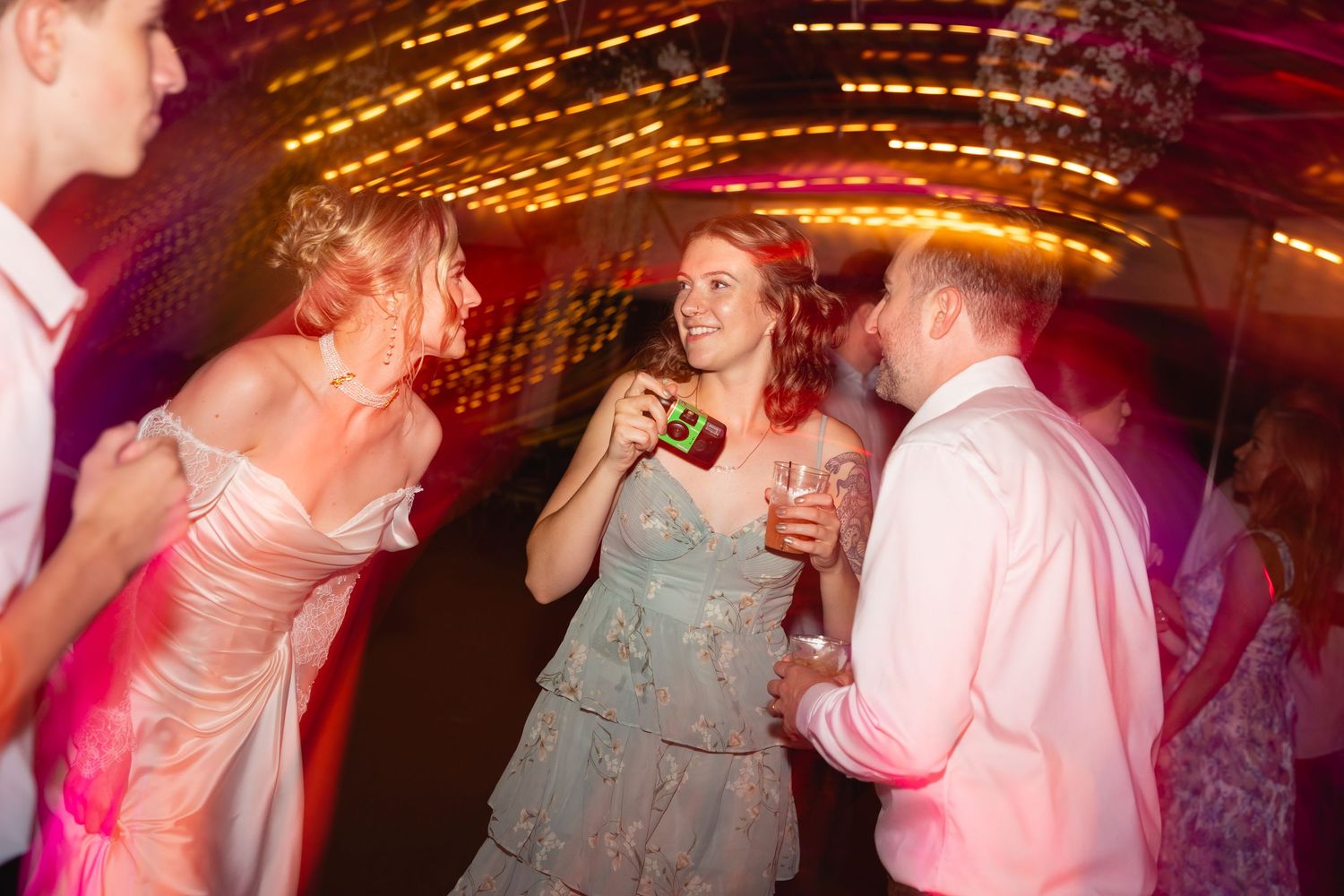 People dancing together at a lively party with colorful pink lighting and string lights overhead.