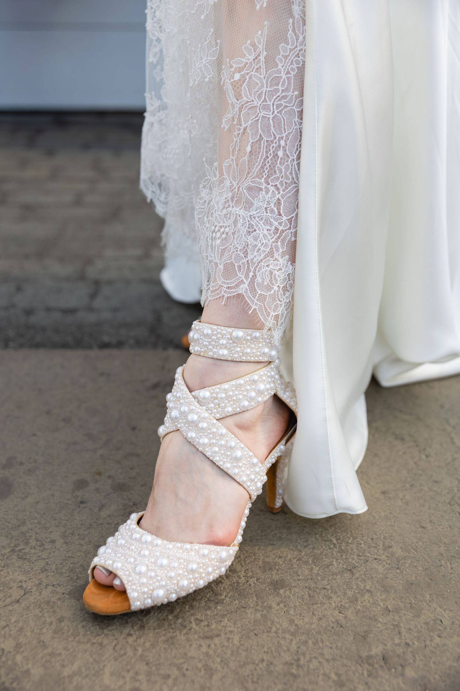 Elegant white lace wedding shoe with pearl details and strappy design shown against concrete floor with dress hem visible.