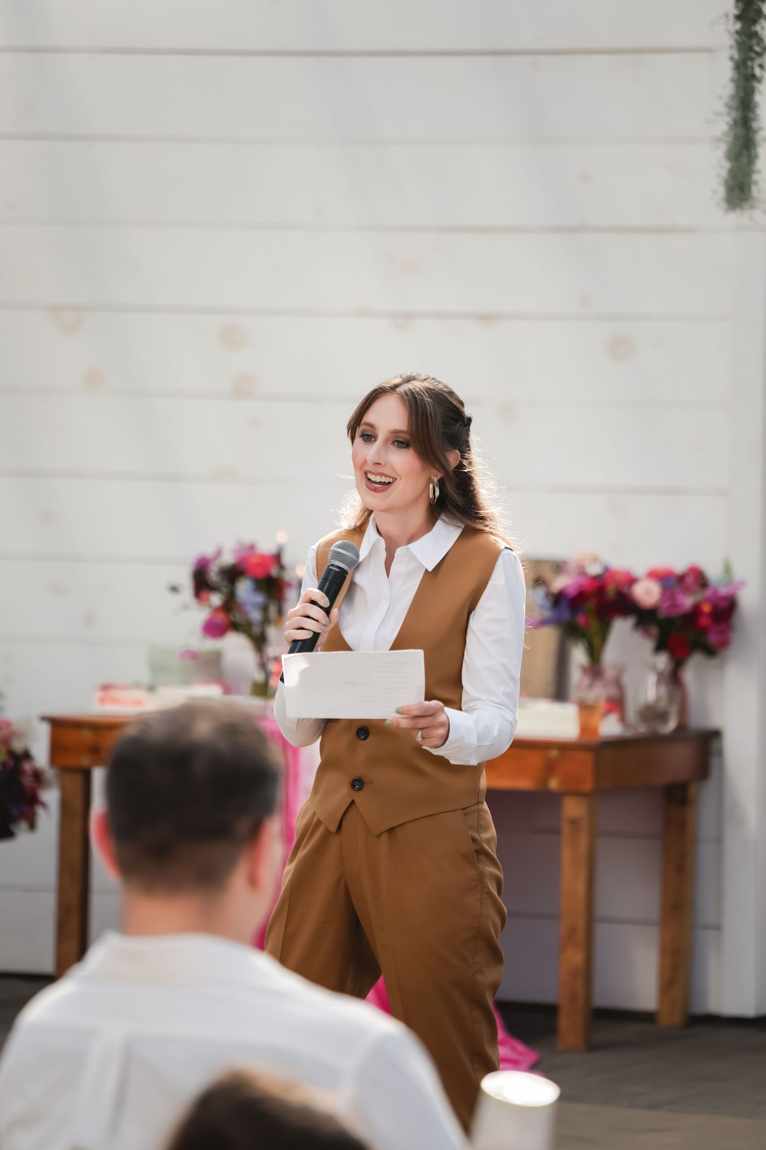 A person in brown attire speaks into a microphone during an indoor event with pink floral decorations.