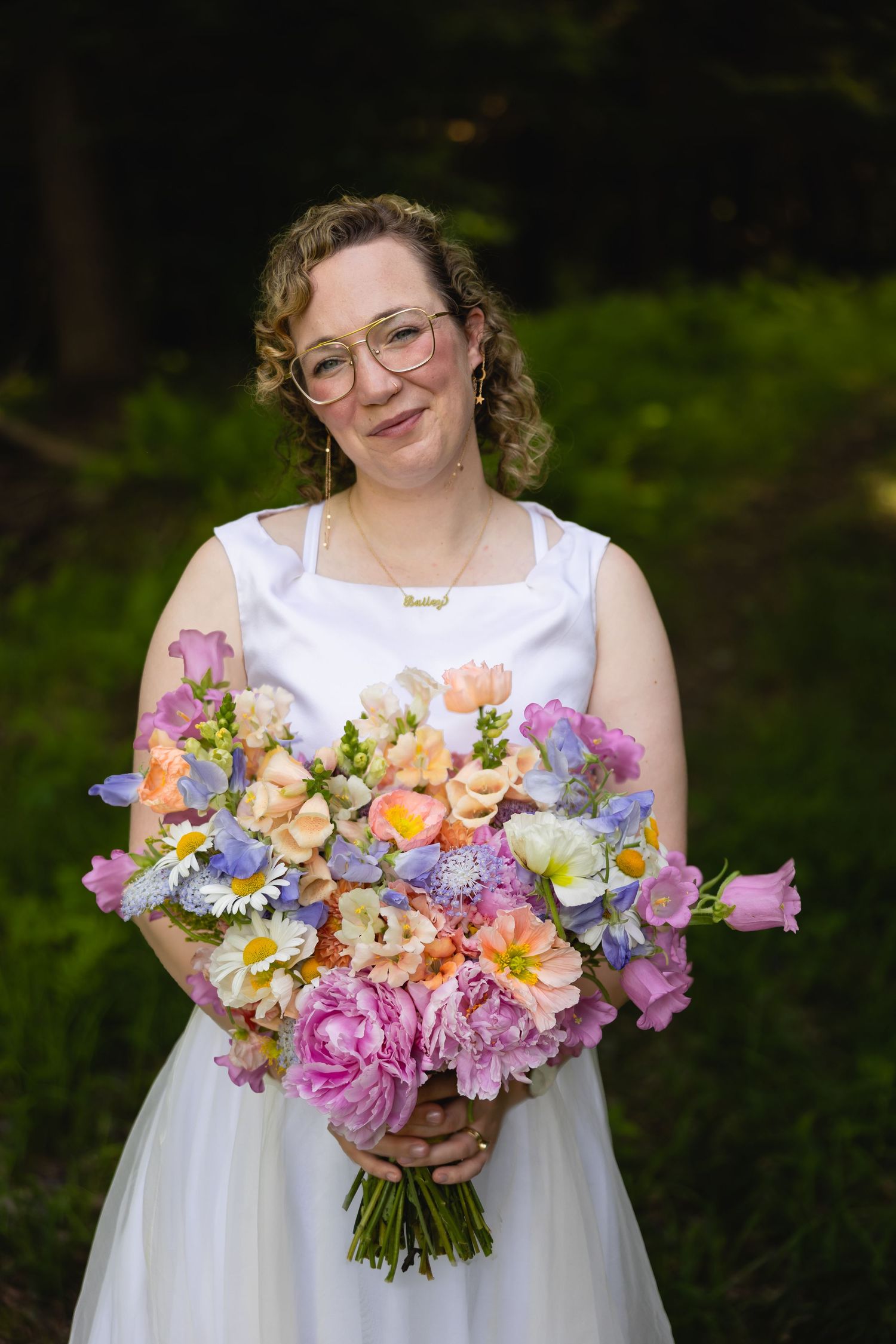 A person in a white dress holds a vibrant spring bouquet with pink, peach and yellow flowers against a green background.