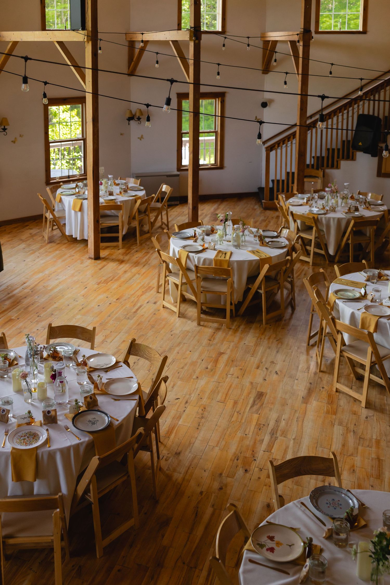 Rustic wedding reception venue with round tables, wooden chairs, and hardwood floors in a bright room with windows.