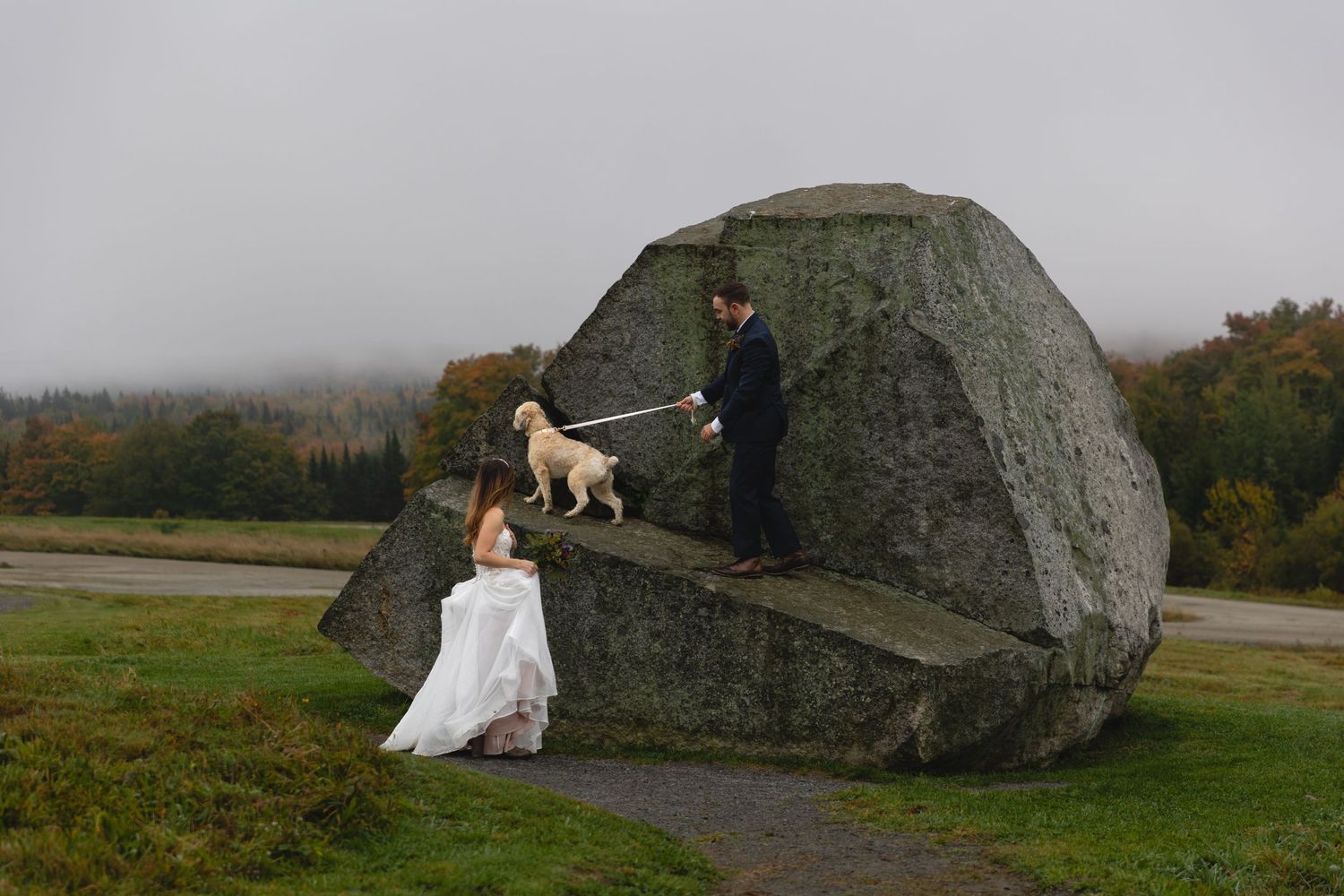 Wedding couple with their dog explore a large boulder formation in a mountain landscape.
