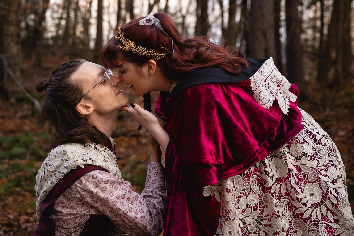 A couple in vintage Victorian-style burgundy and lace clothing share an intimate moment in a forest setting.