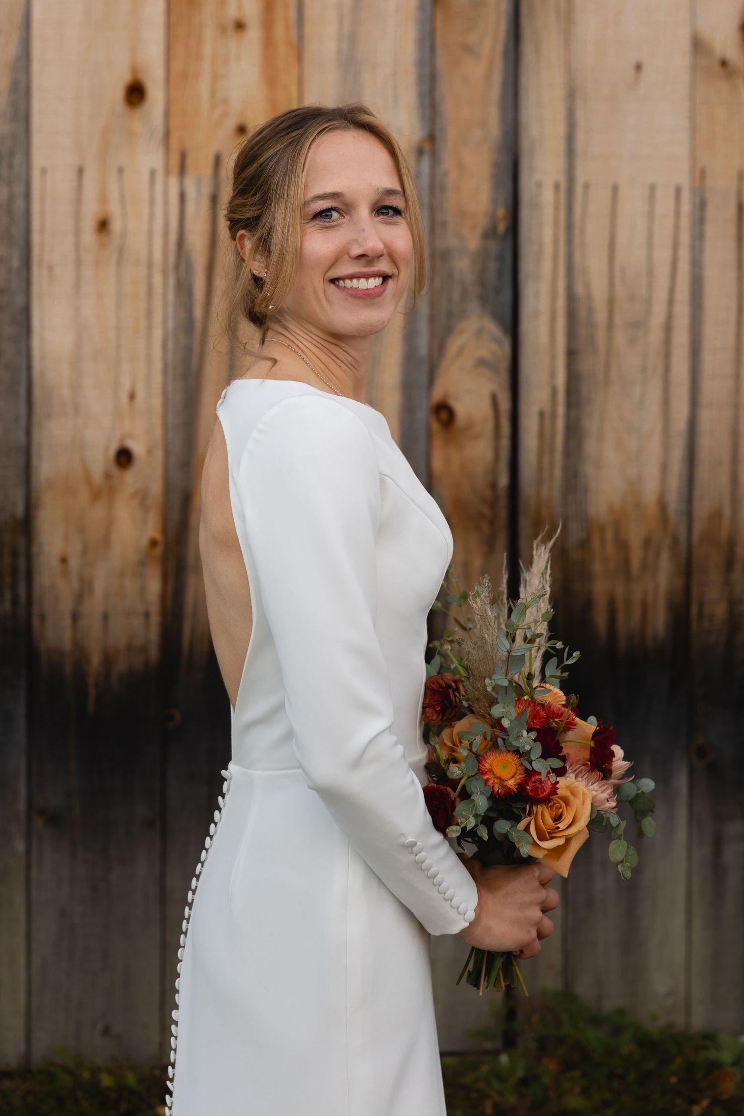 A bride in a simple long-sleeved white dress holds an autumn-colored bouquet against a rustic wooden fence background.