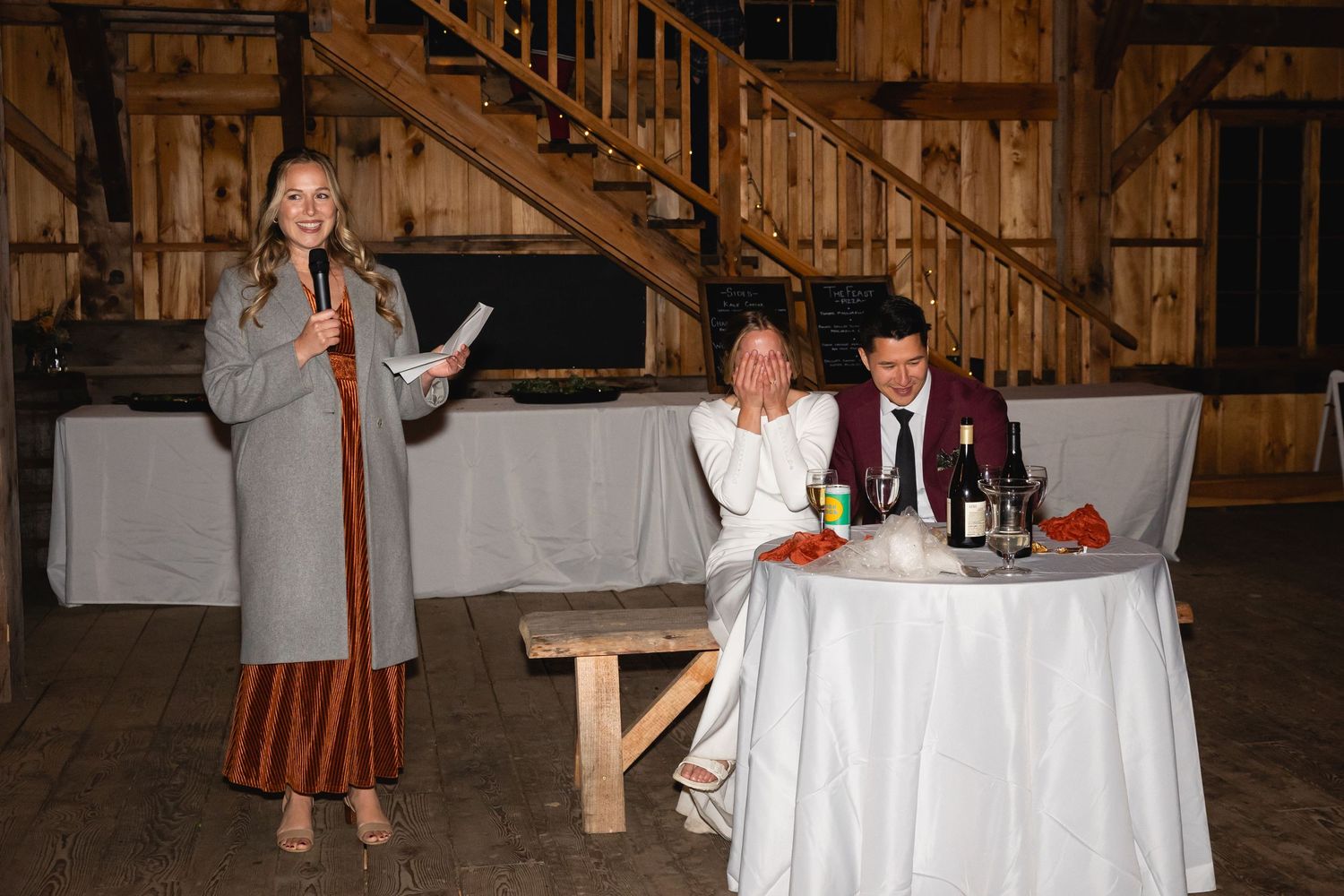Wedding guests celebrate at a rustic barn reception with a speaker standing near a white-clothed table.
