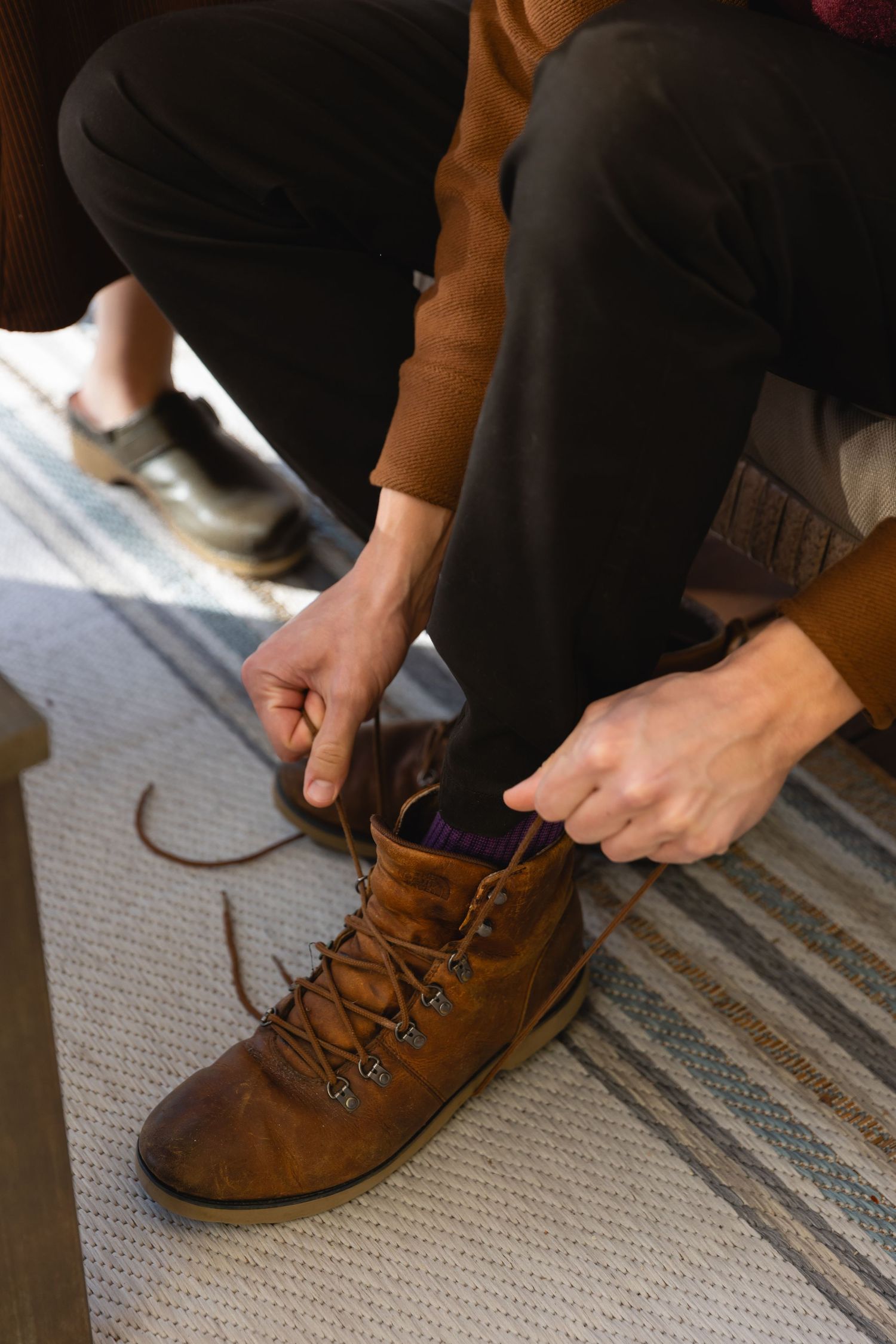 Someone ties the laces of a brown leather ankle boot while sitting on a striped rug.