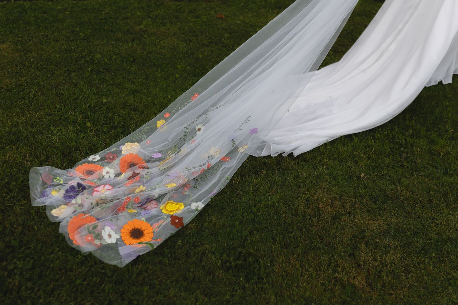 White wedding veil adorned with colorful floral embellishments trailing on grass.