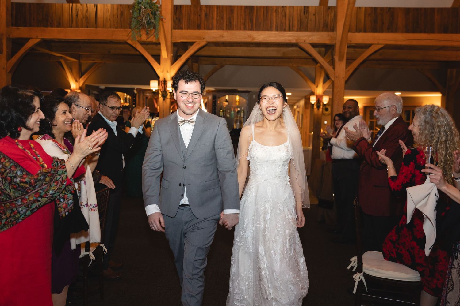 Newlyweds walk down aisle in rustic barn venue while wedding guests cheer and applaud.