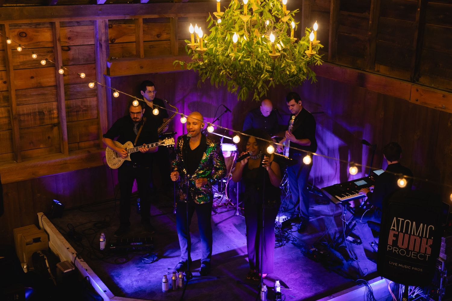 A band performs on a dimly lit stage with purple lighting and a gold chandelier overhead in a rustic venue.