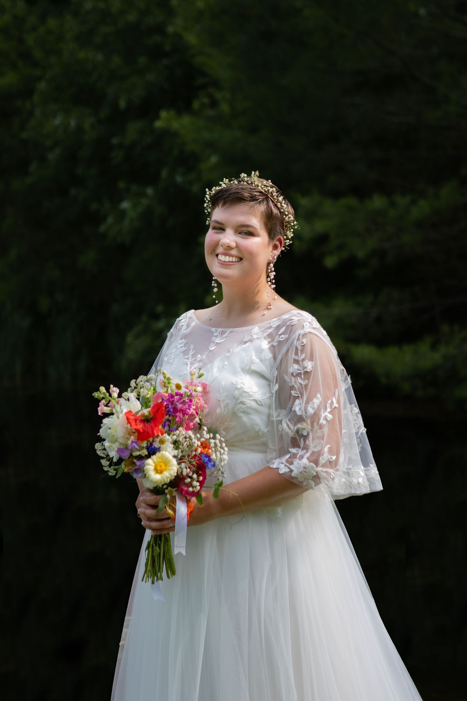 Bride in lace-sleeved white gown holds colorful bouquet against forest backdrop.