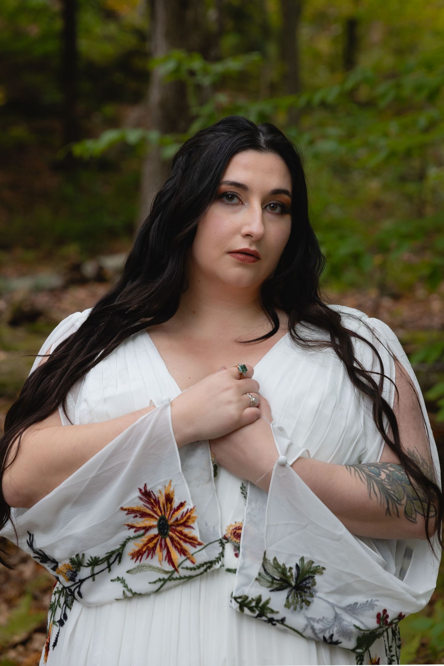 A bride in a flowing white dress with floral embroidery poses against an autumn forest backdrop.