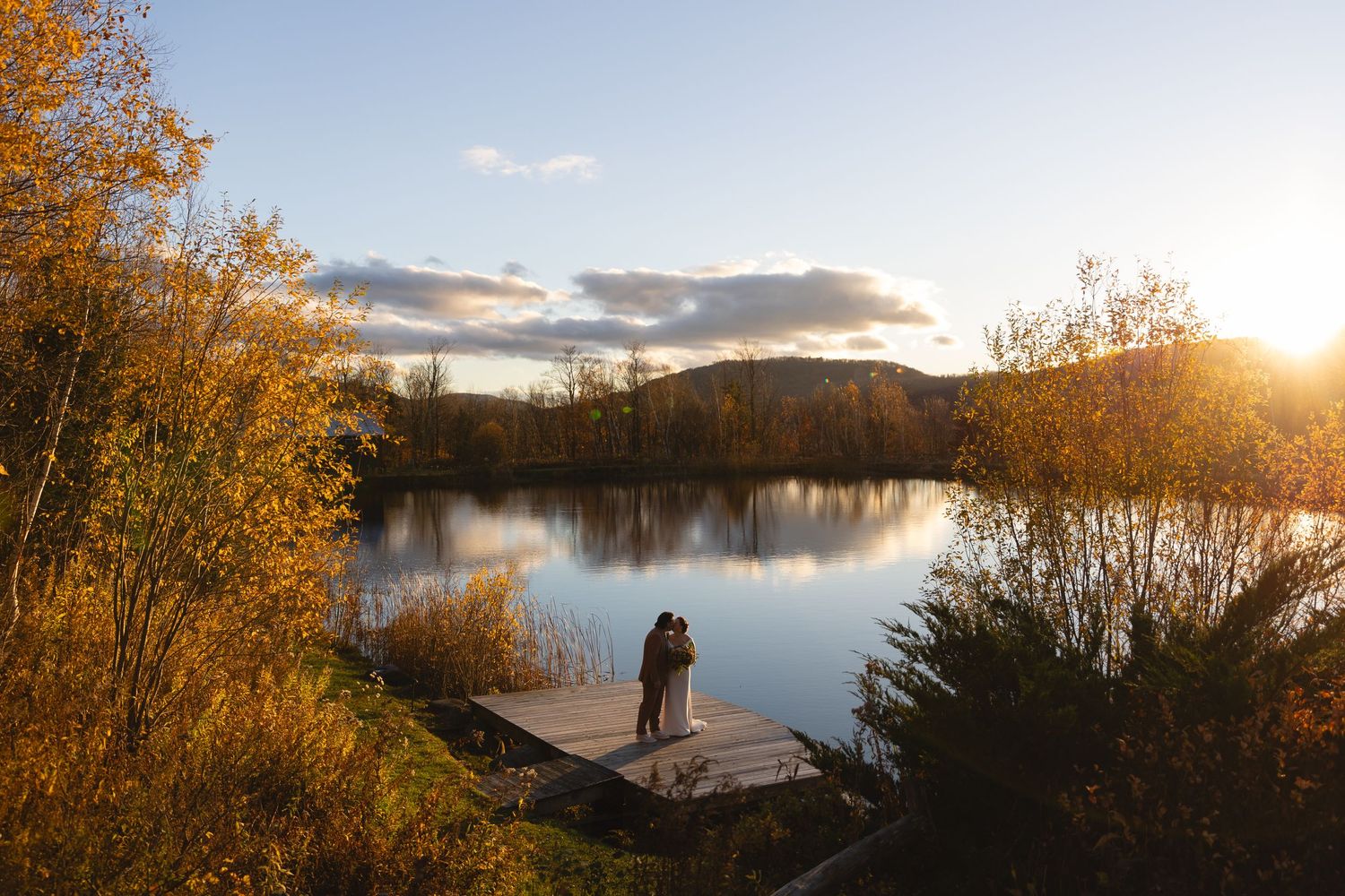 Couple silhouetted on a wooden dock at sunset overlooking a calm lake surrounded by autumn foliage.