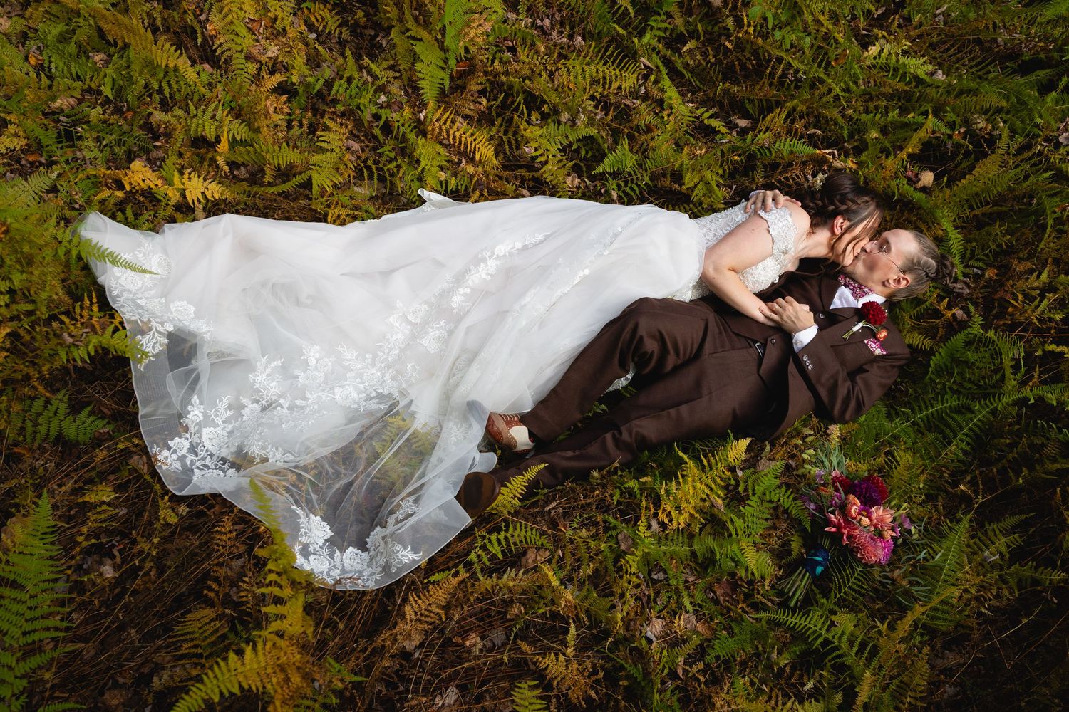 Bride and groom lie together in golden ferns, with her white lace gown and bouquet spread around them.