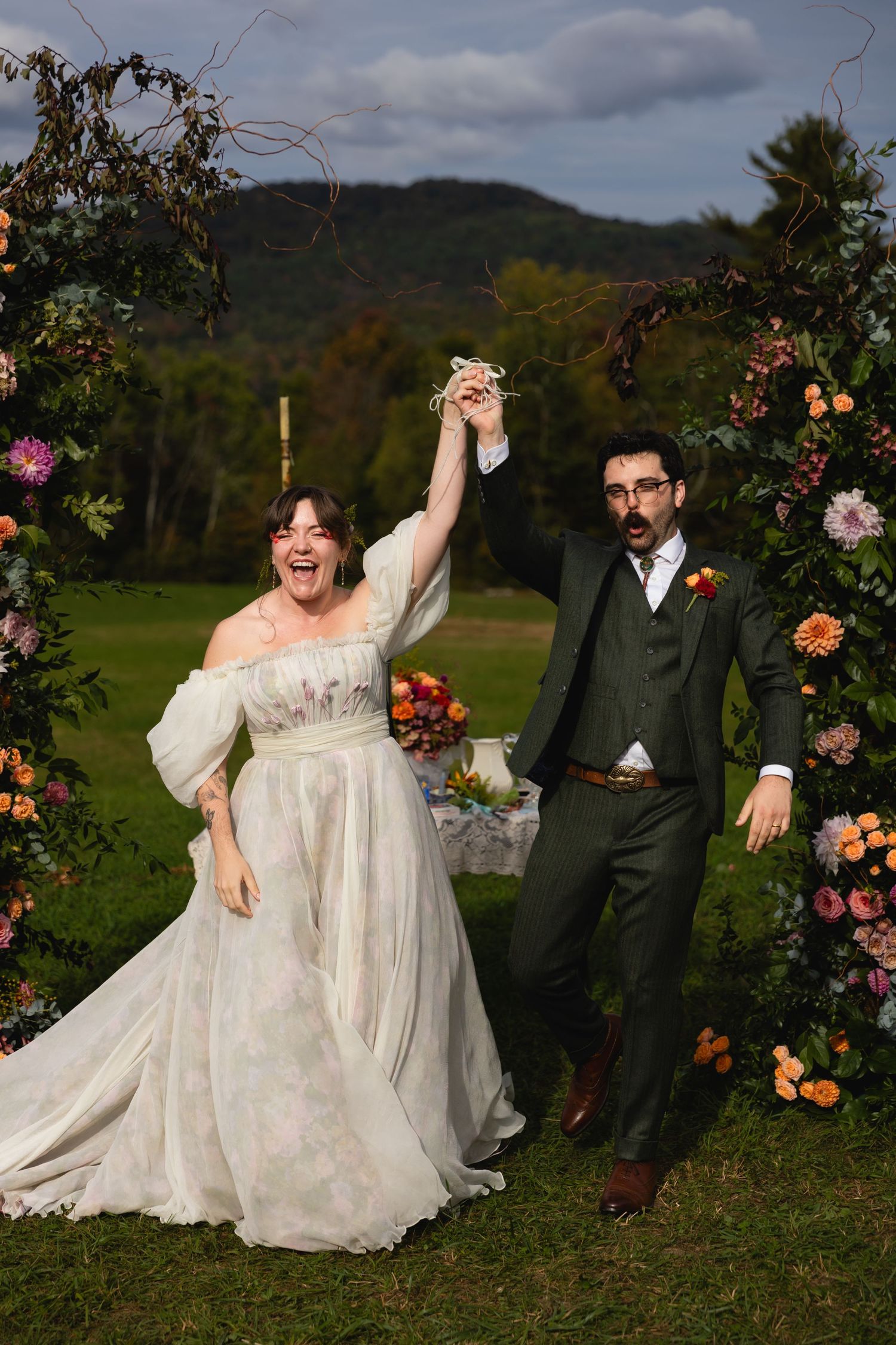 A jubilant bride and groom raise joined hands beneath a floral arch with mountain views behind them.