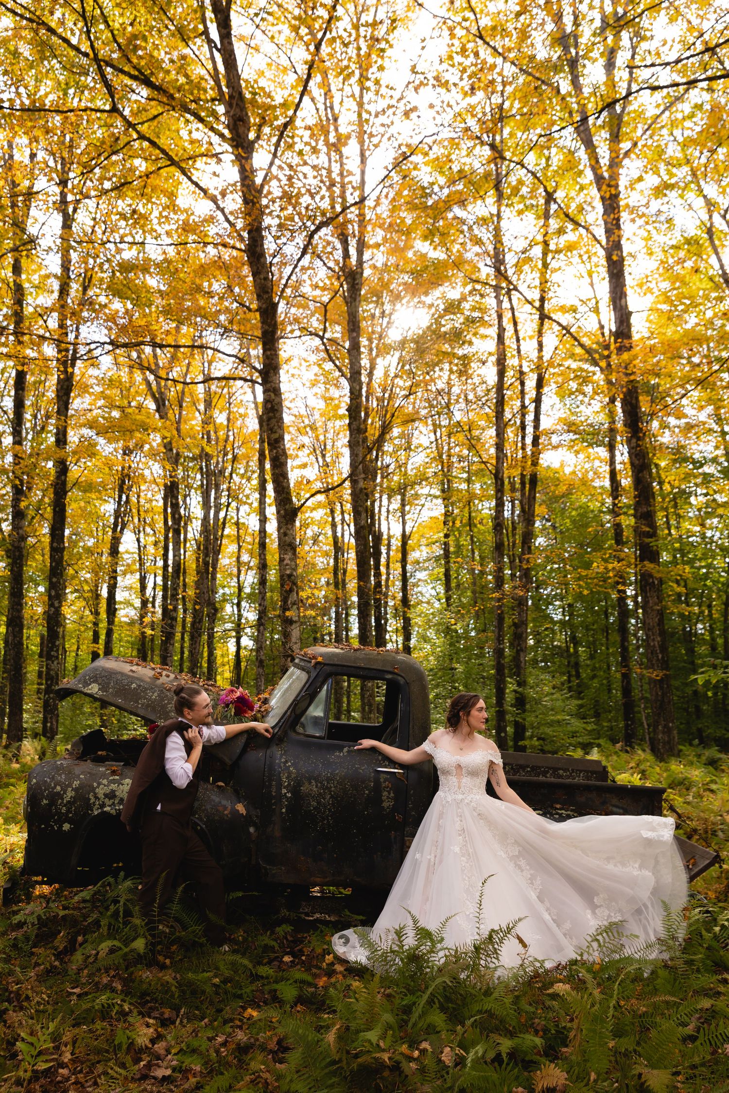 Bride and groom pose beside a rustic vintage truck surrounded by golden fall foliage.