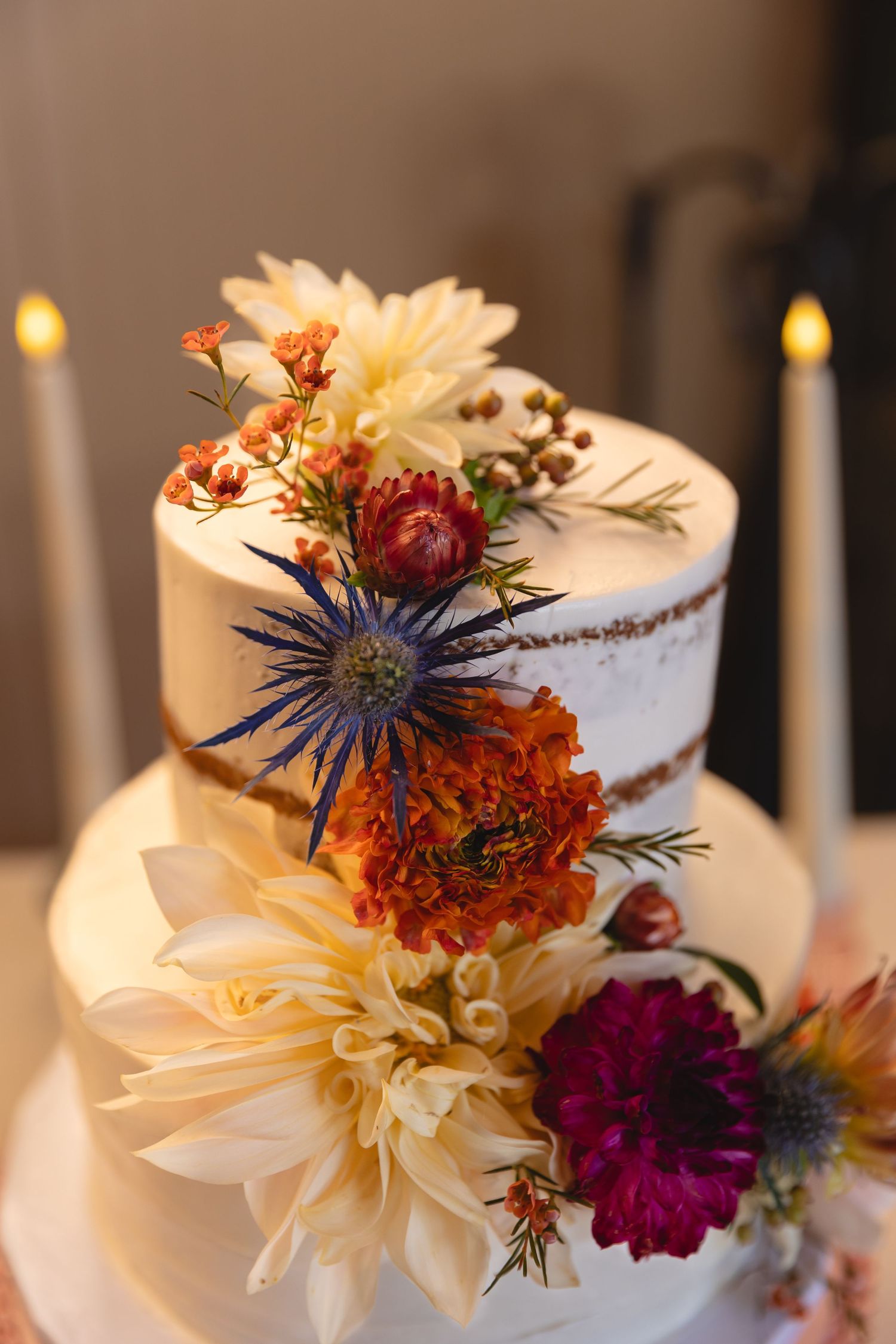 Rustic wedding cake decorated with colorful autumn flowers including dahlias, thistles and marigolds against a candlelit background.