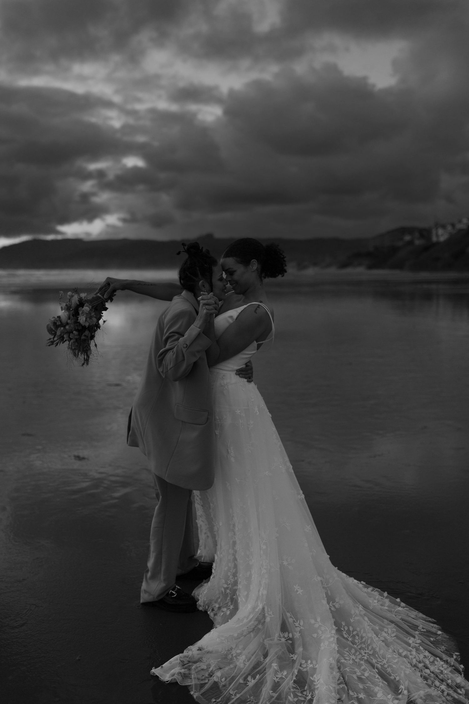 A black and white beach wedding photo with dramatic storm clouds and mountain scenery.