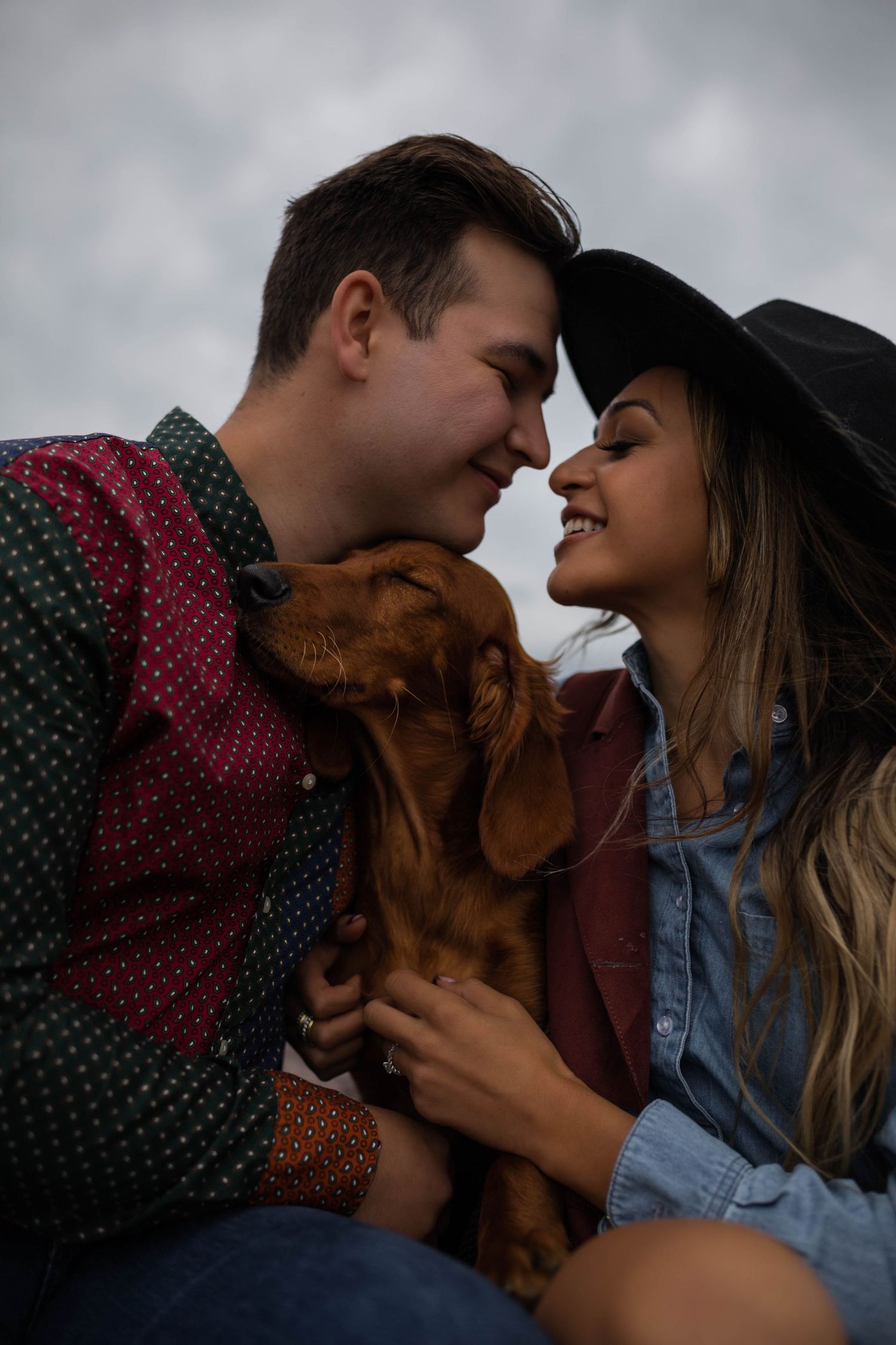 A joyful couple shares an intimate moment with their brown dog on a cloudy day.
