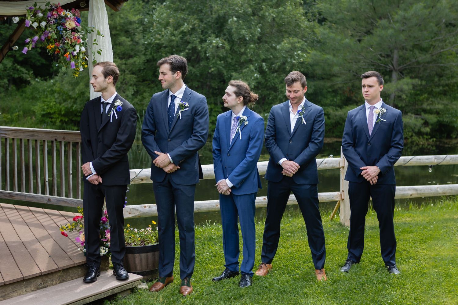 Groomsmen in navy and black suits standing on wooden deck during outdoor wedding.