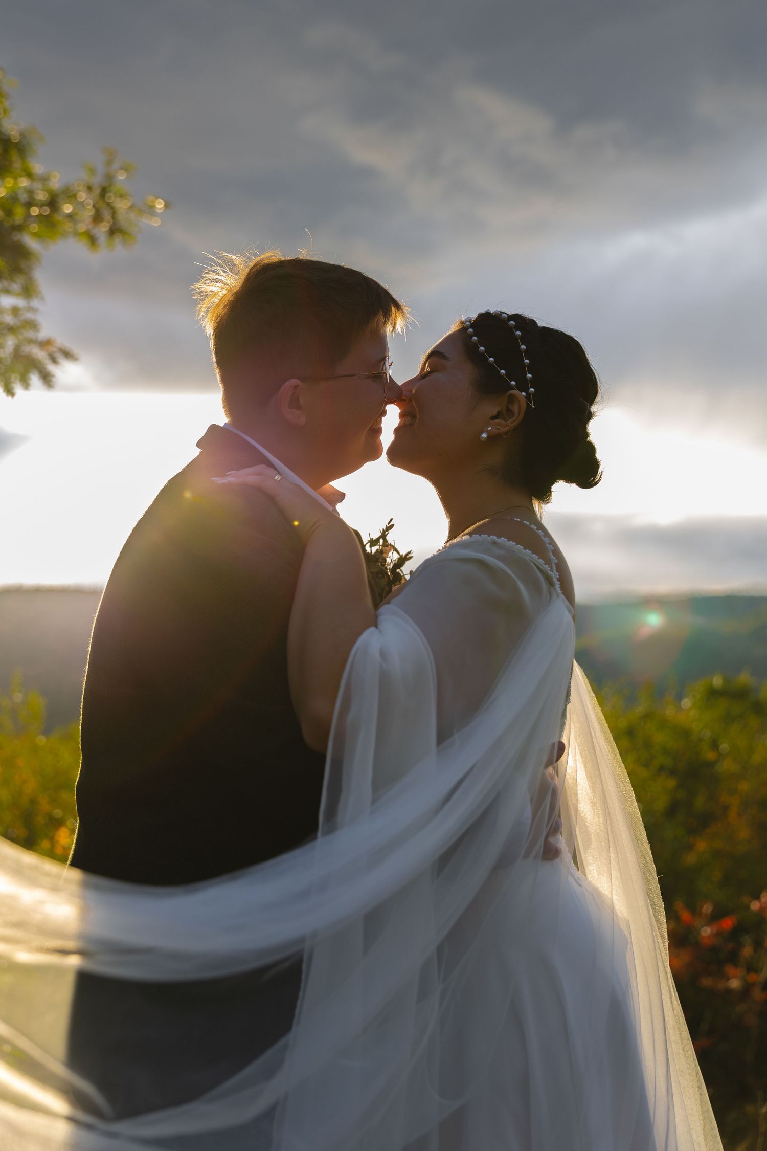 Sunlit wedding portrait with flowing veil and dramatic sky behind the kissing couple.
