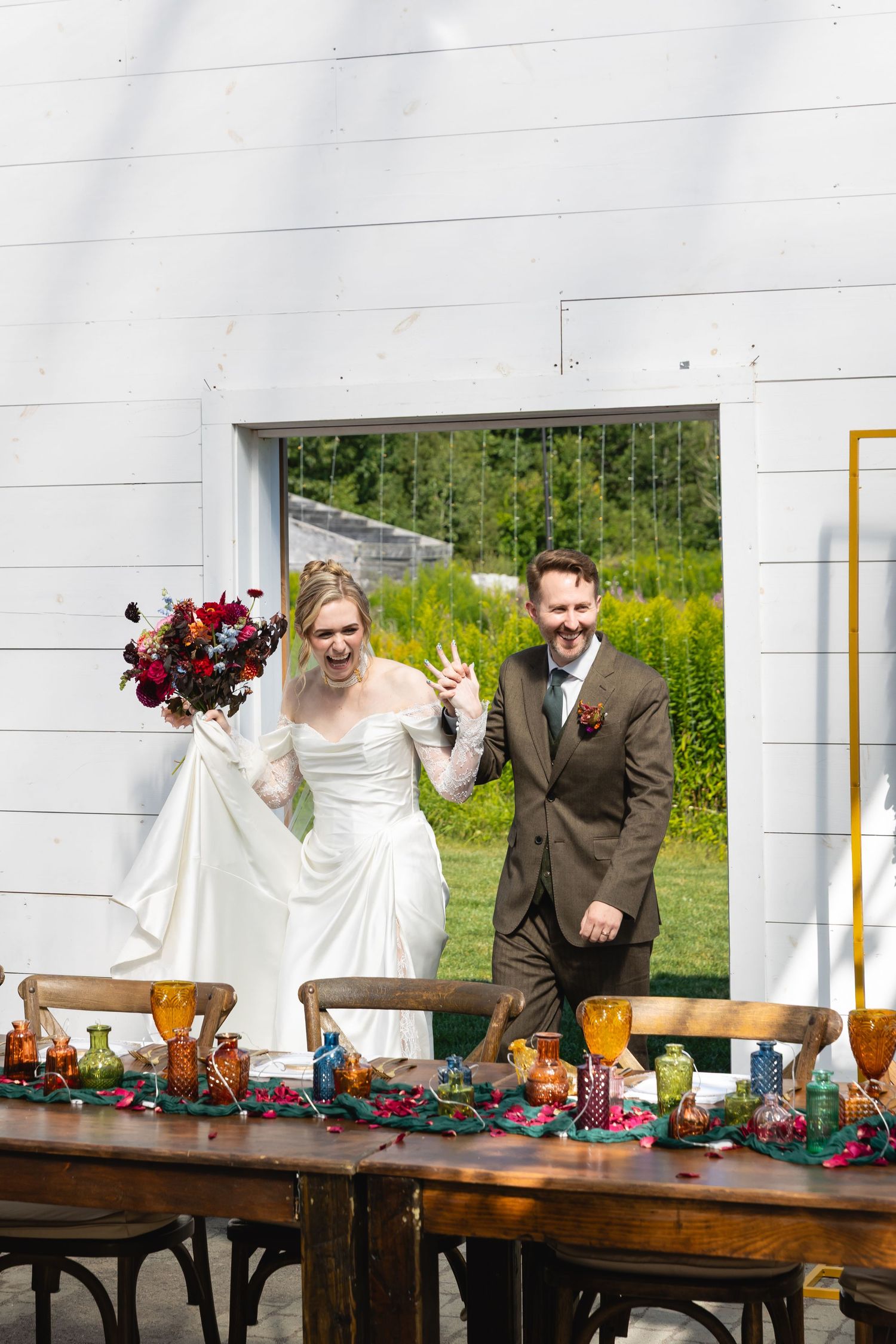 A couple celebrates at their wedding reception next to a rustic wooden table decorated with amber glasses and floral arrangements.